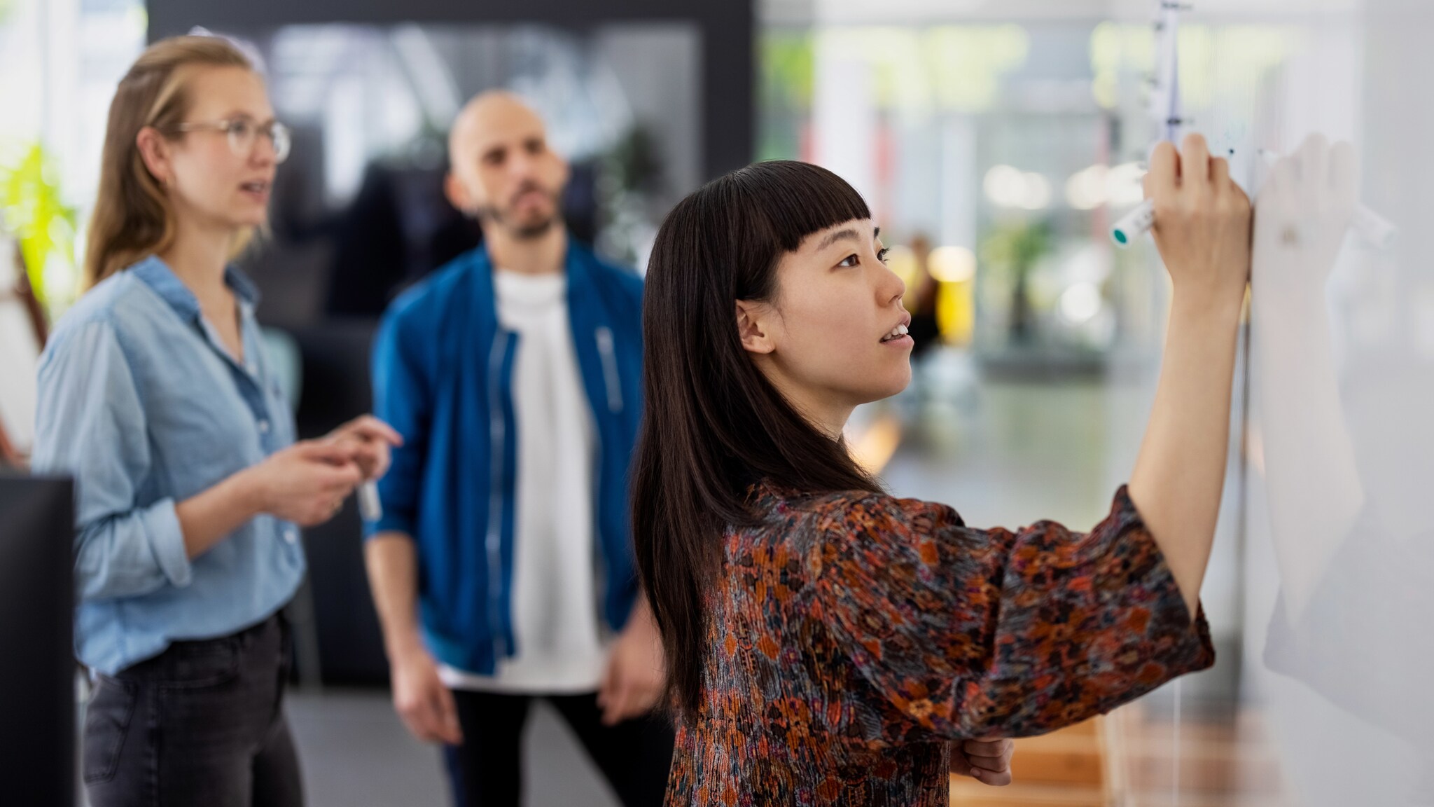 Eine Frau schreibt mit einem Marker auf ein Whiteboard, dahinter stehen zwei Personen, die ihr dabei zuschauen.