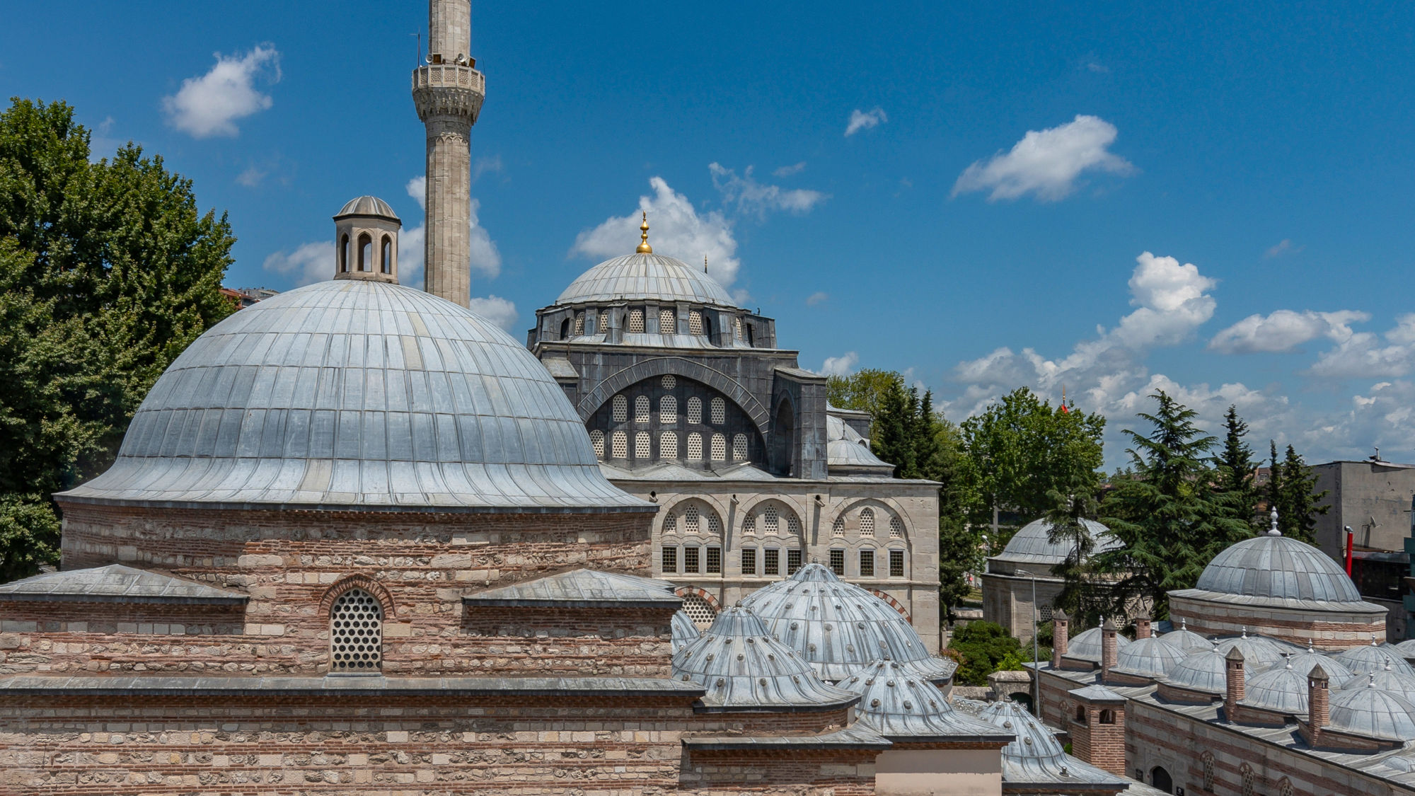 Dach eines türkischen Bads mit unterschiedlichen Kuppeln vor einer Moschee unter blauem Himmel.