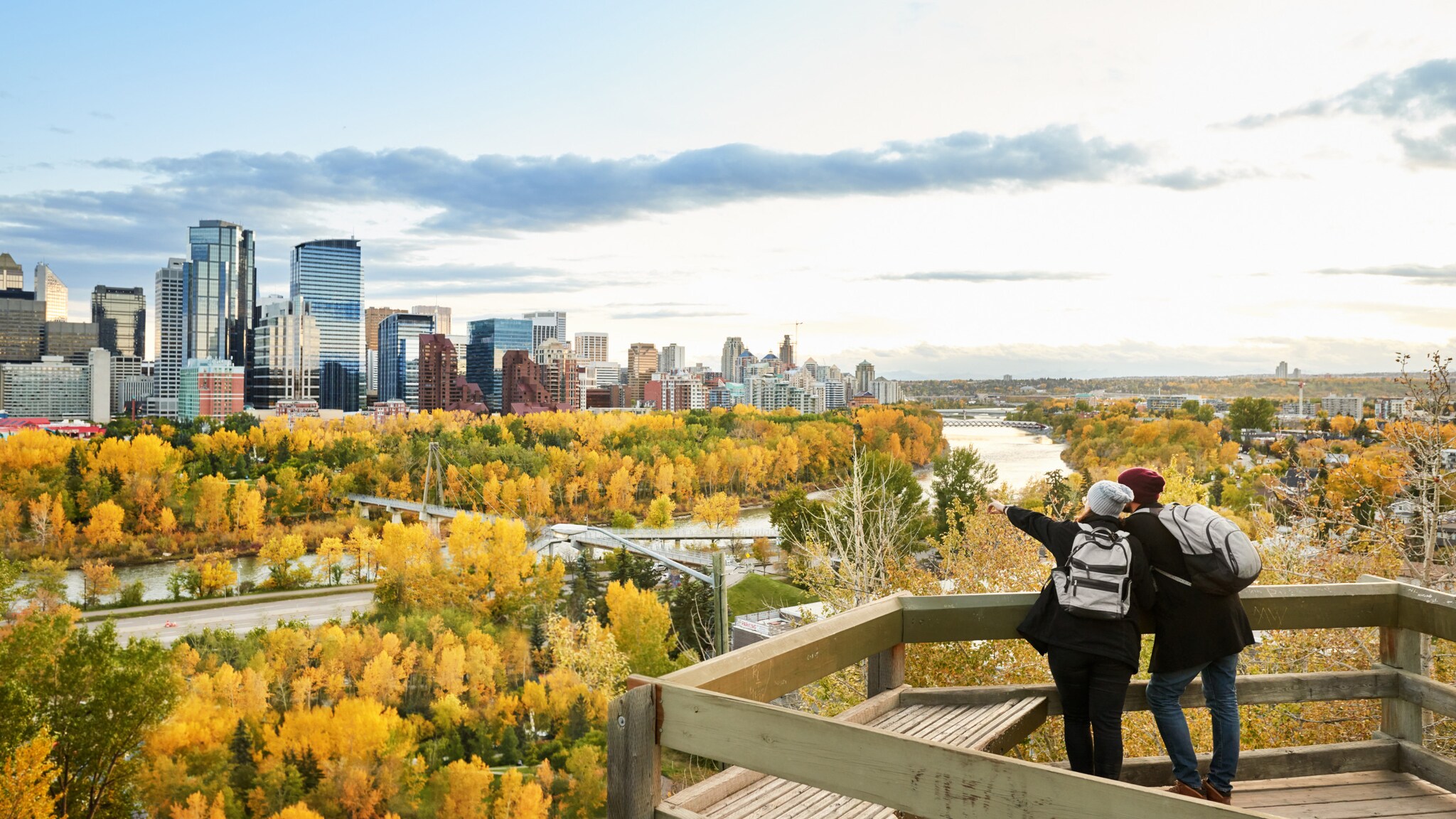 Zwei Personen mit Rucksäcken stehen auf einer Aussichtsplattform und blicken auf die Stadt Calgary mit herbstlich gefärbtem Wald und Hochhäusern. Zwei Personen mit Rucksäcken stehen auf einer Aussichtsplattform und blicken auf die Stadt Calgary mit herbstlich gefärbtem Wald und Hochhäusern.