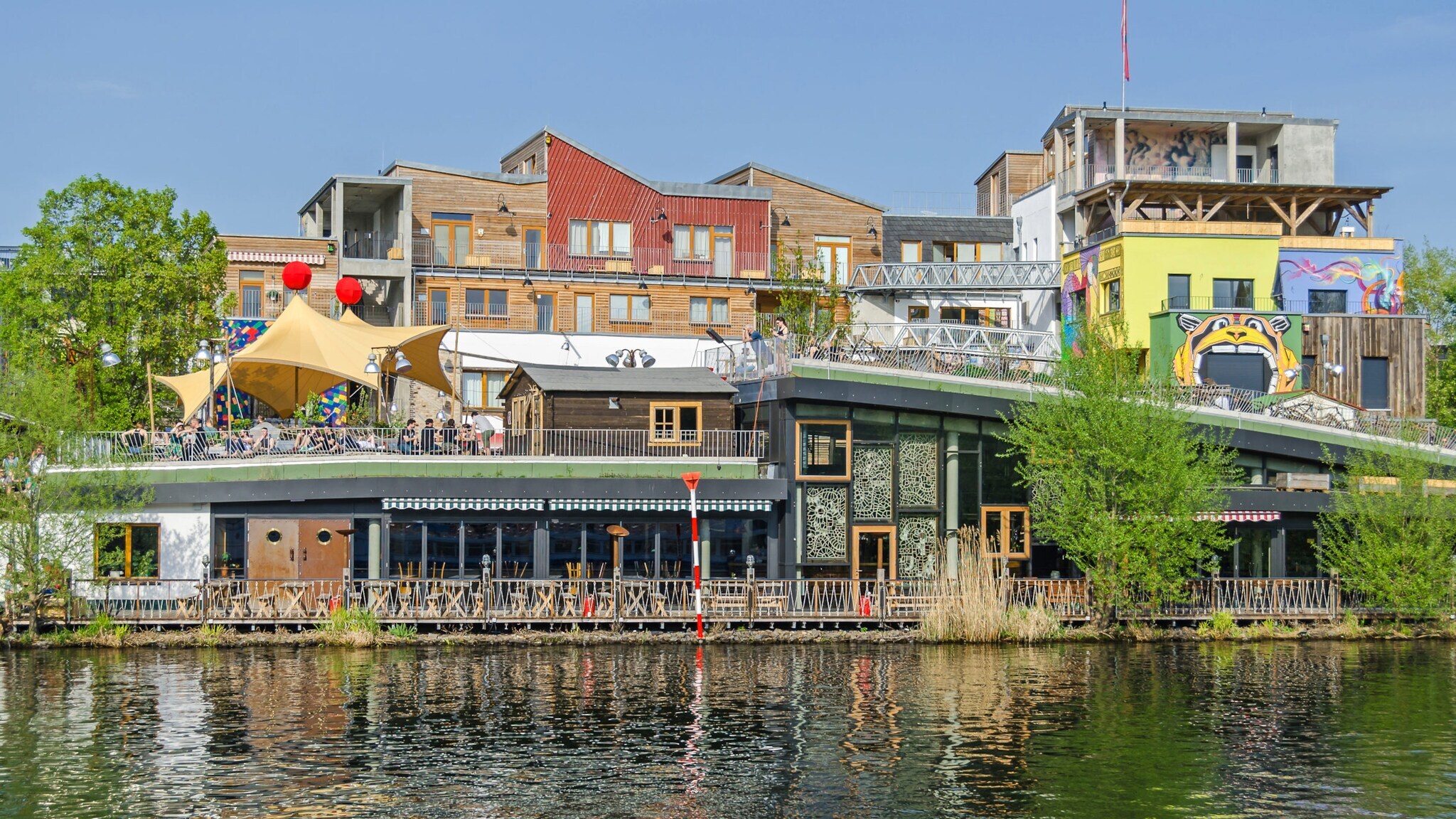 Bunte Gebäude und Cafés am Ufer eines Kanals mit Menschen auf der Terrasse und Spiegelung im Wasser