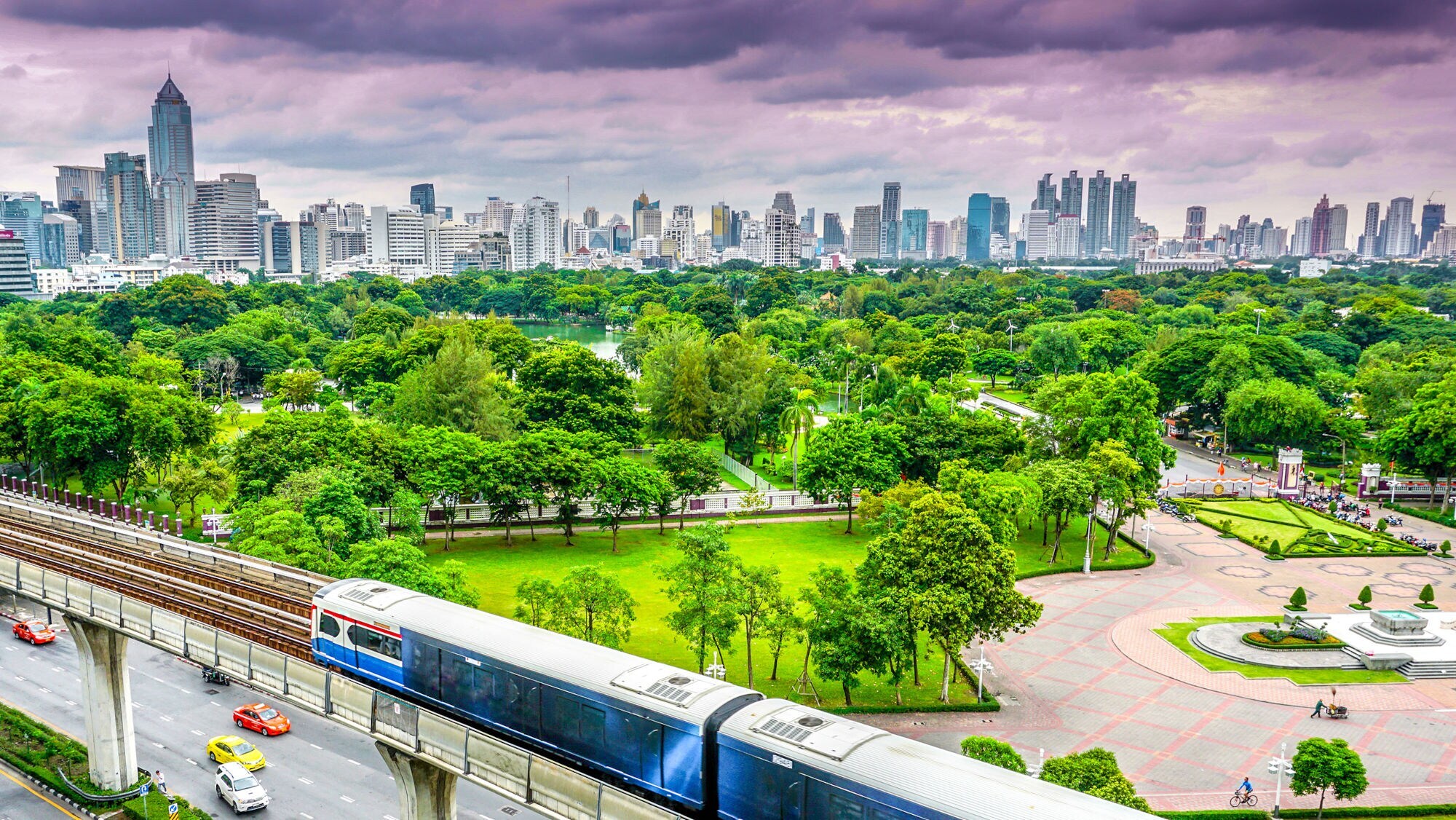 Vor einer Skyline mit Hochhäusern befindet sich ein grüner Park mit Bäumen und einer Wiese, davor fährt eine Straßenbahn.