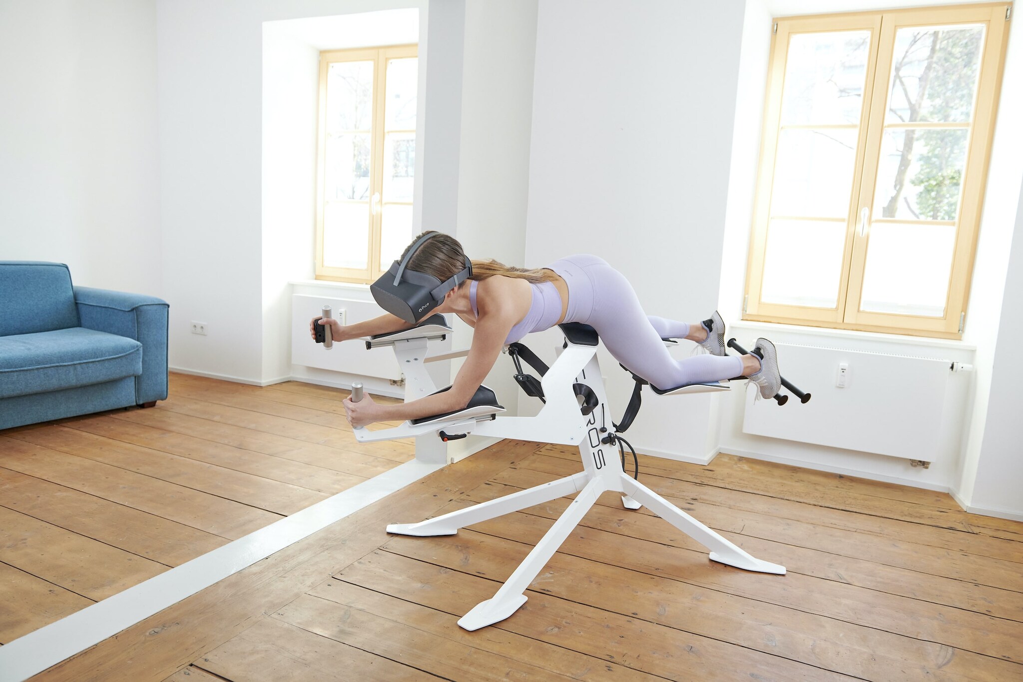 Eine Frau in fliederfarbenem Sport-Outfit liegt auf einem weißen Heimtrainer, der wie ein Flugsimulator aussieht und trägt eine virtuelle Brille