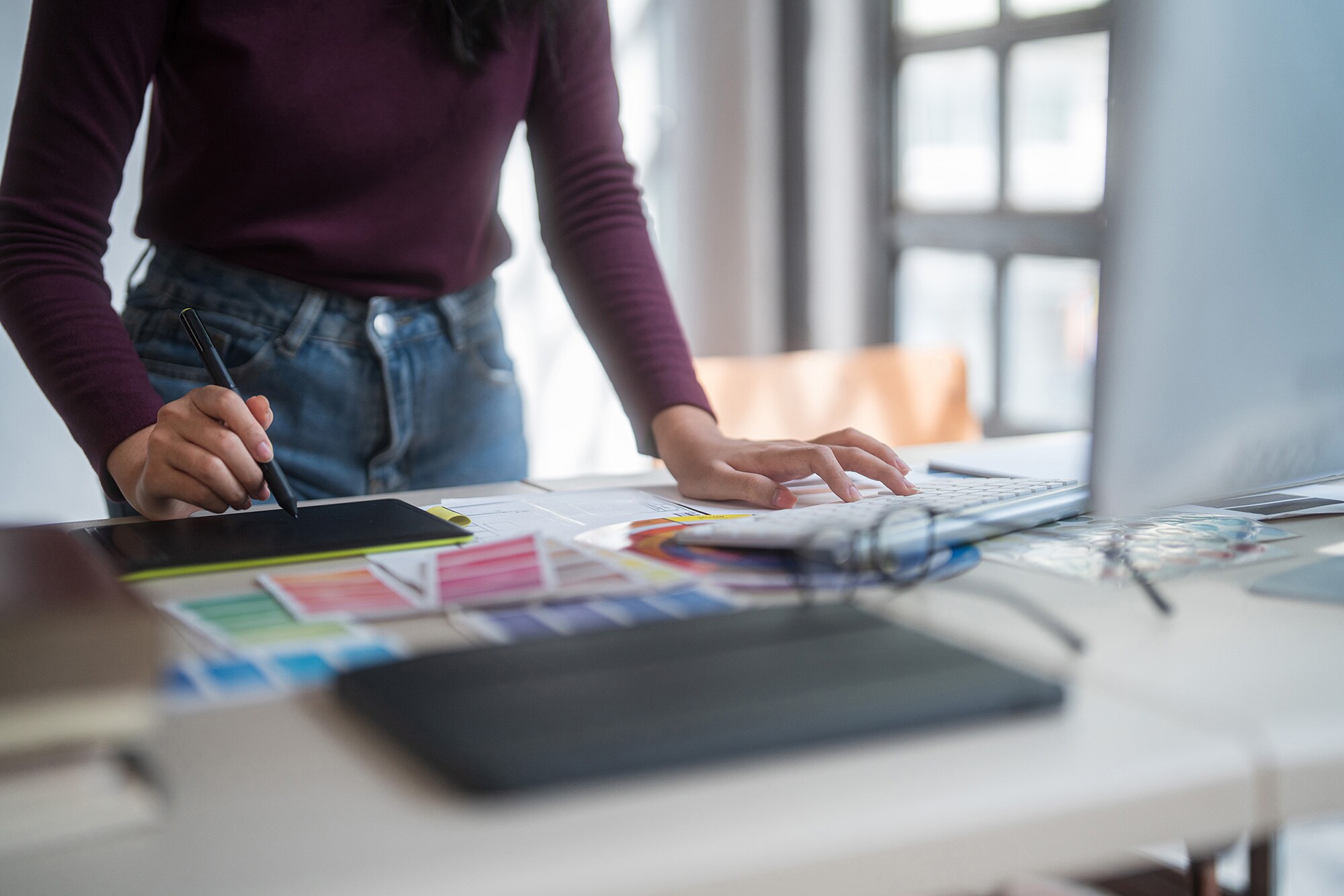 Eine Frau steht an ihrem Schreibtisch und arbeitet am Computer sowie am Tablet. Brandbook Basics: Schritt für Schritt