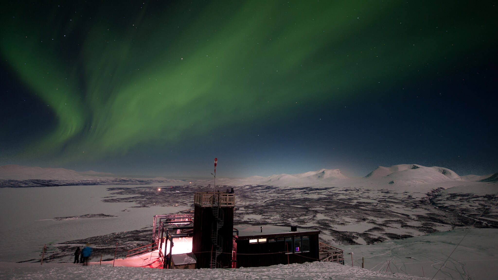 Grünes Nordlicht am Nachthimmel über einer verschneiten Landschaft mit einem kleinen, beleuchteten Gebäude und zwei Personen in Winterkleidung. Grünes Nordlicht am Nachthimmel über einer verschneiten Landschaft mit einem kleinen, beleuchteten Gebäude und zwei Personen in Winterkleidung.