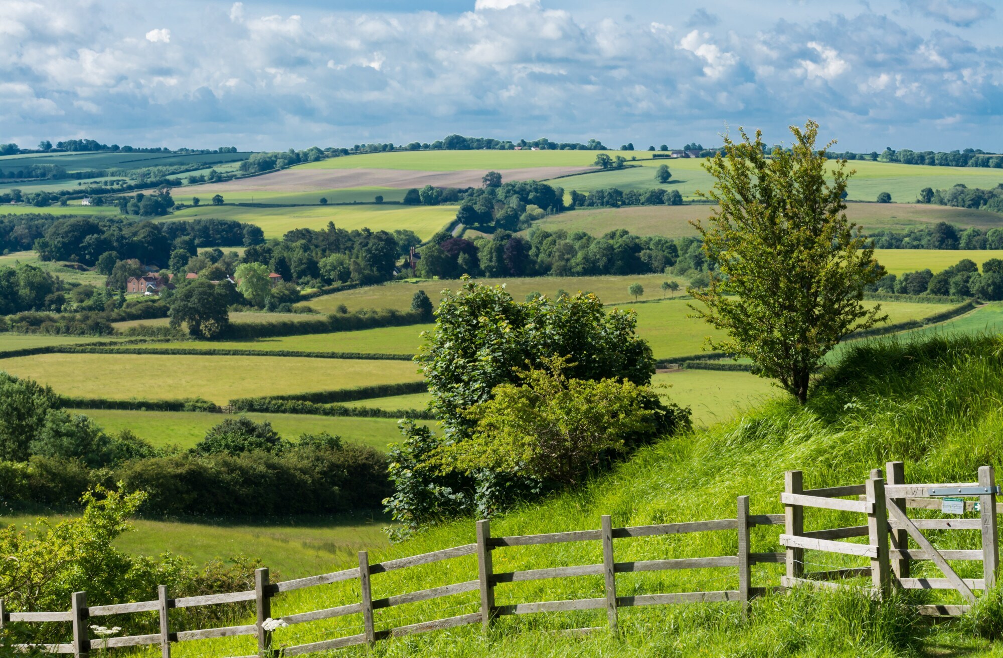 Grüne Hügellandschaft mit landwirtschaftlichen Parzellen, im Vordergrund ein Holzzaun. Grüne Hügellandschaft mit landwirtschaftlichen Parzellen, im Vordergrund ein Holzzaun.