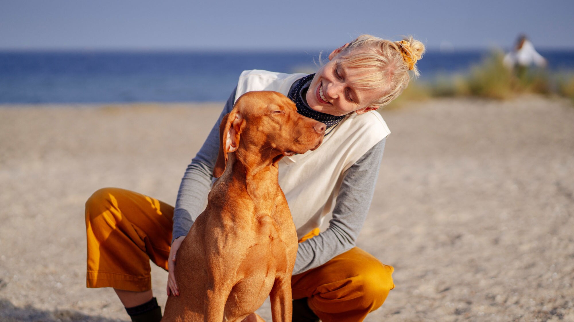 Eine Frau kniet hinter einem großen Hund mit goldfarbenem Fell und schaut ihn lächelnd an auf einem breiten Sandstrand bei Sonnenschein.