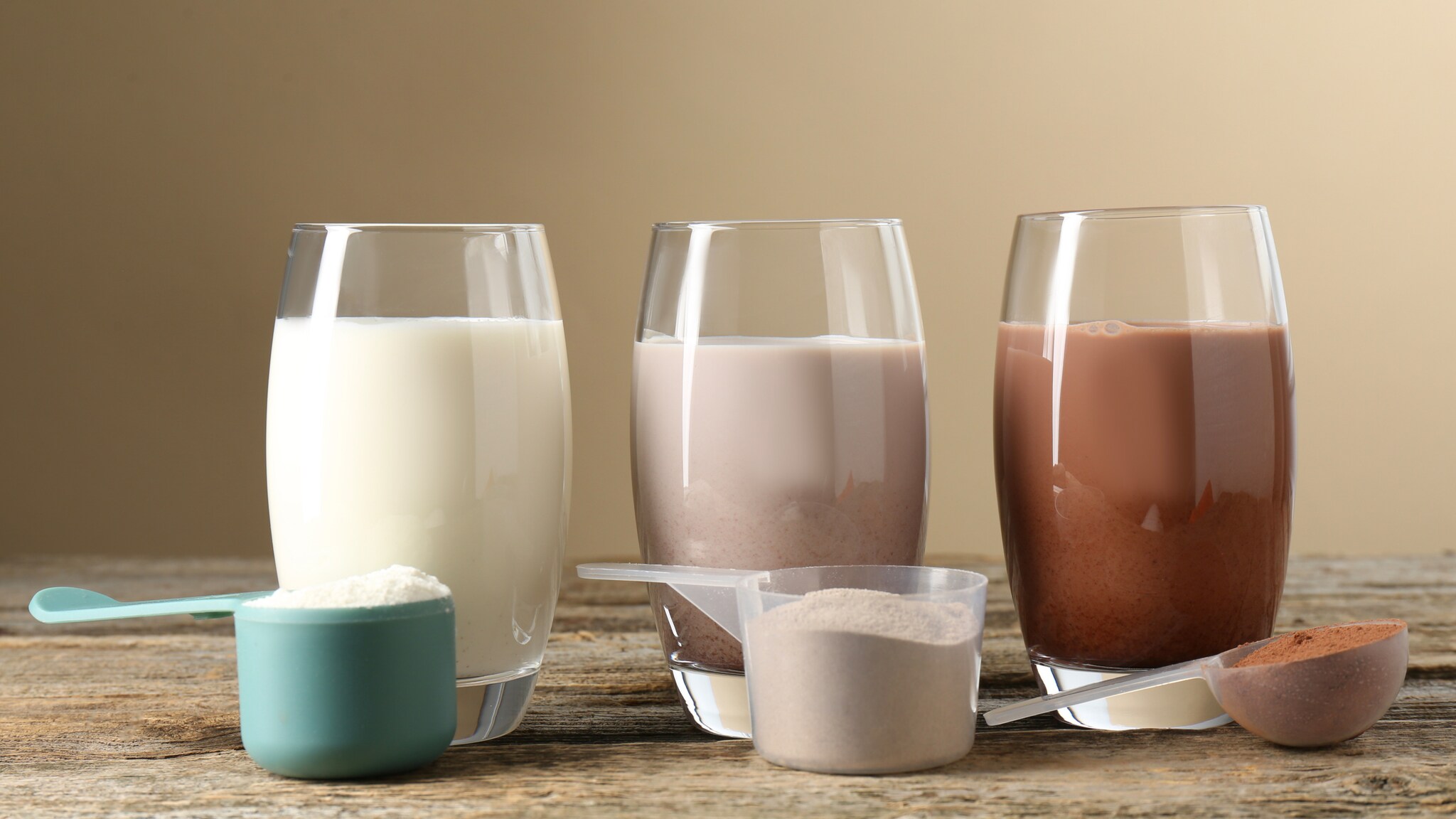 Drei Gläser mit weißem, hellbraunem und dunklem Proteinshake, davor drei Löffel mit weißem, hellbraunem und dunklem Proteinpulver auf Holztisch Drei Gläser mit weißem, hellbraunem und dunklem Proteinshake, davor drei Löffel mit weißem, hellbraunem und dunklem Proteinpulver auf Holztisch