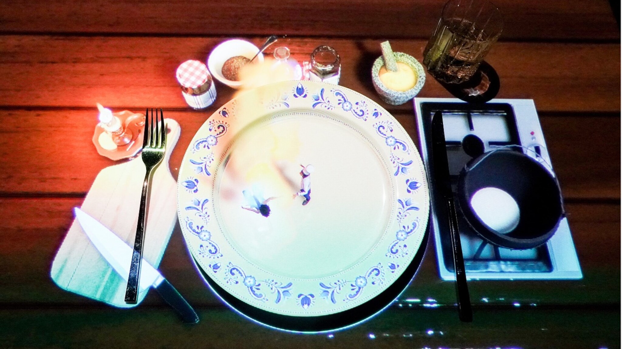 Ein gedeckter Tisch mit einem großen weißen Teller, auf dem zwei kleine Figuren stehen. Daneben befinden sich ein Messer, eine Gabel, ein Glas, Gewürzgläser und eine Pfanne mit einem Ei.