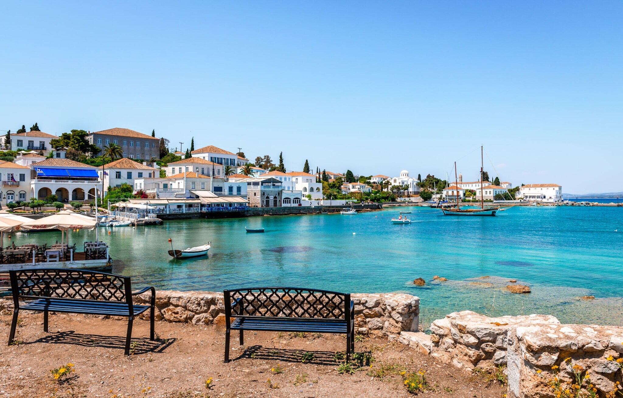 Kleiner Hafenort mit einzelnen Fischerbooten im türkisblauen Wasser, im Vordergrund zwei Sitzbänke am Ufer. Kleiner Hafenort mit einzelnen Fischerbooten im türkisblauen Wasser, im Vordergrund zwei Sitzbänke am Ufer.