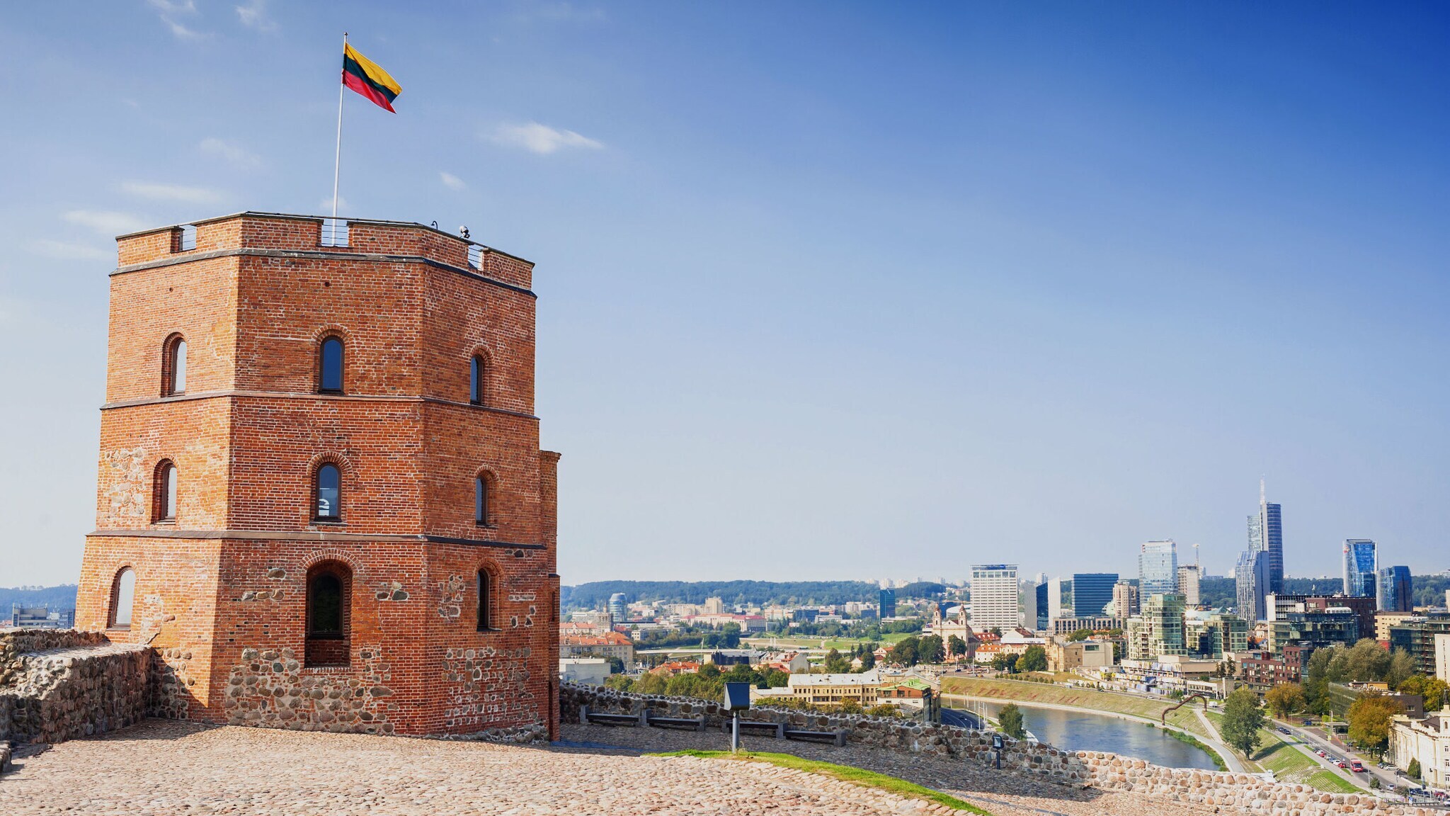 Eckiger Burgturm aus rotem Backstein mit litauischer Flagge auf einer Festung auf einem Hügel vor Stadtpanorama.
