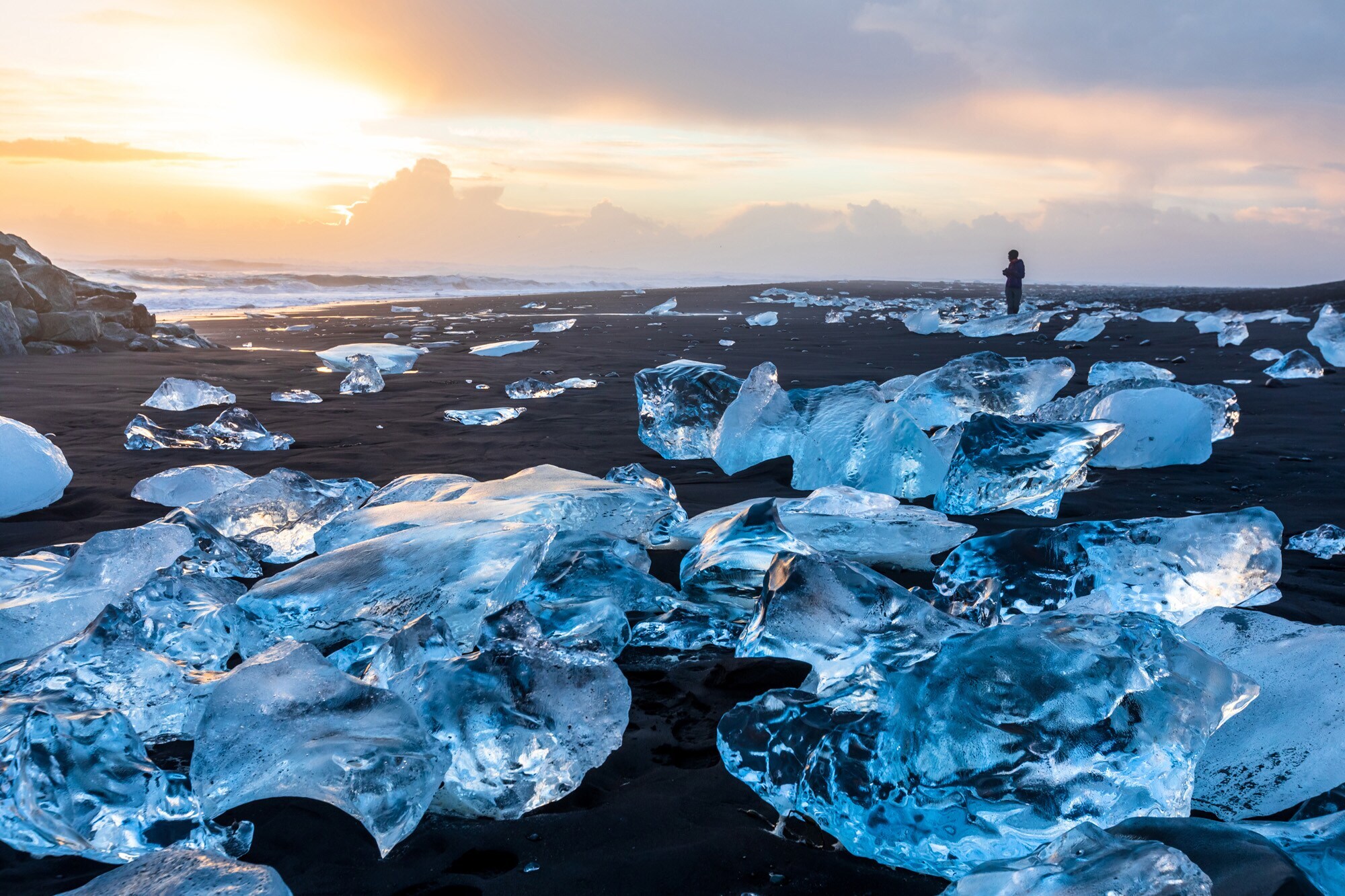 Eine Person steht am Diamond Beach in Island und blickt auf blaue Eisbrocken, die auf dem schwarzen Sand liegen und im Licht der aufgehenden Sonne glitzern