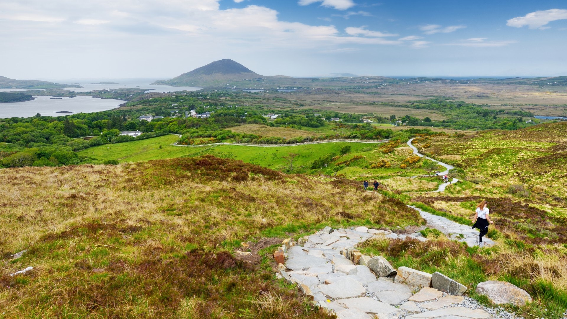 wandern-in-irland-landschaft Heide- und Wiesenlandschaft, durch die sich ein steiniger Weg mit wandernden Personen darauf schlängelt, im Hintergrund ein Berg und Wasser.