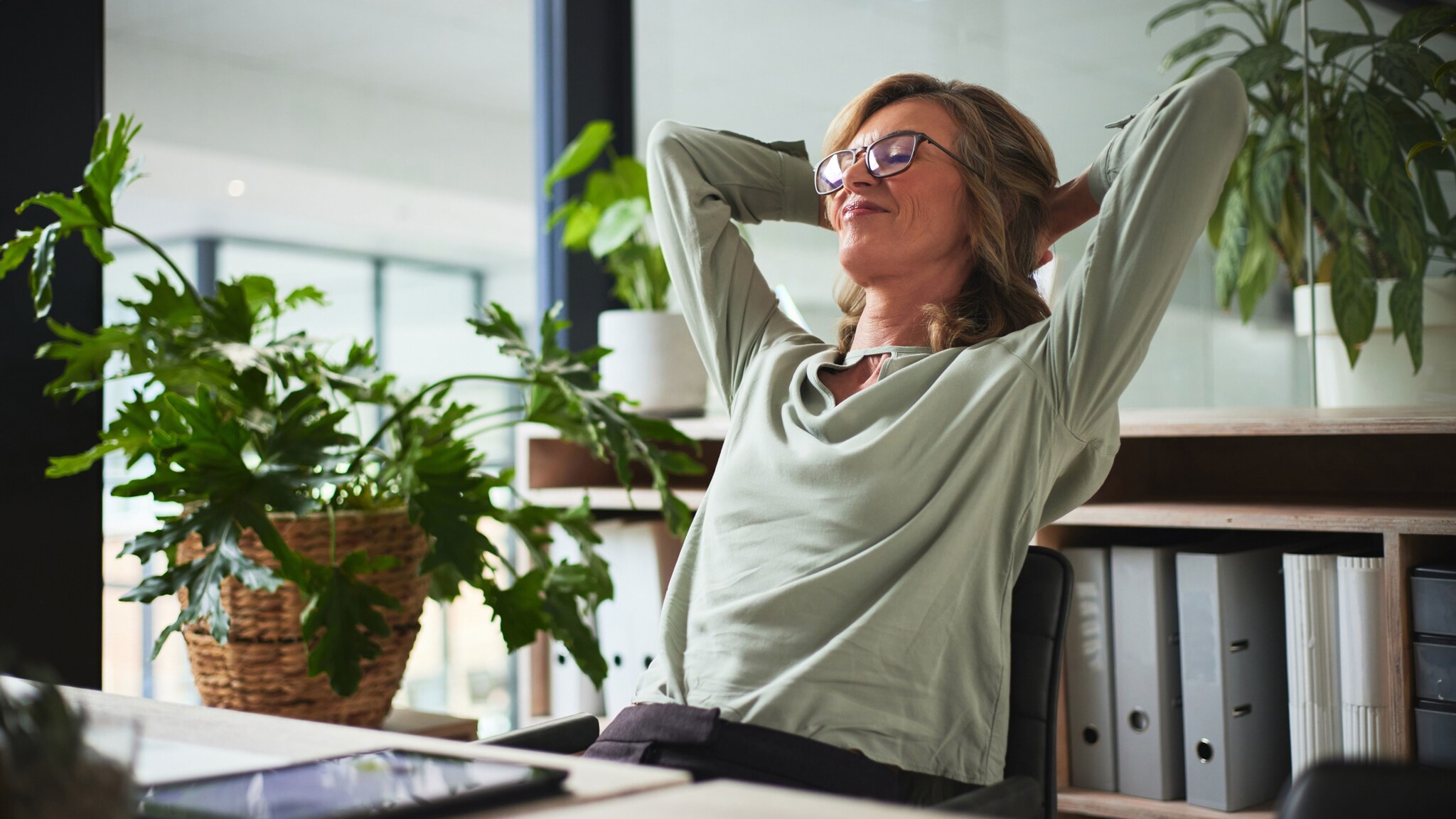 Eine Frau lehnt mit hinter dem Kopf verschränkten Armen in einem Schreibtischstuhl in einem Raum mit Pflanzen und mit Ordnern in Regalen.