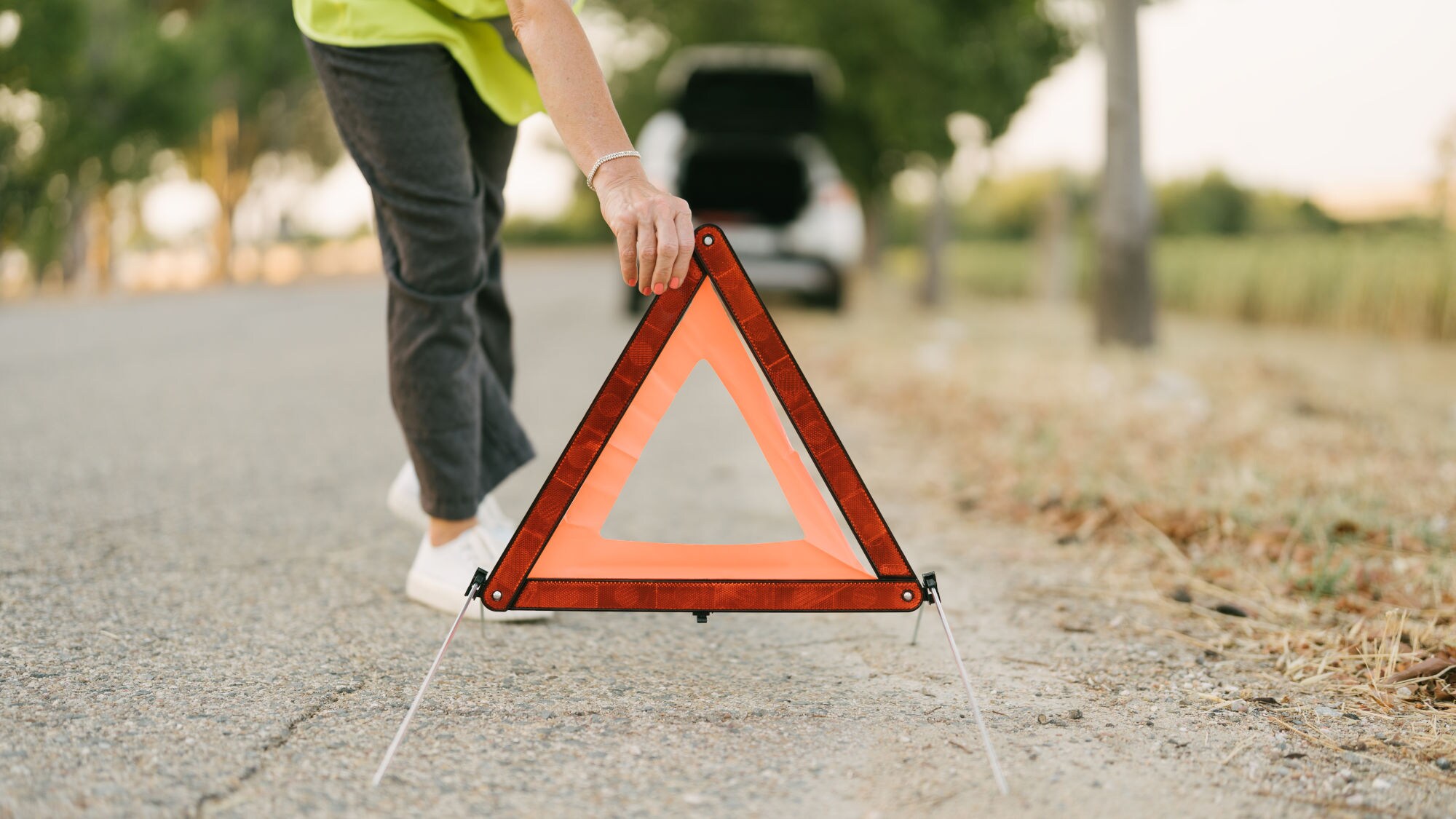 Person stellt ein rotes, dreieckiges Warndreieck mit reflektierendem Rand auf eine asphaltierte Straße. Person stellt ein rotes, dreieckiges Warndreieck mit reflektierendem Rand auf eine asphaltierte Straße.