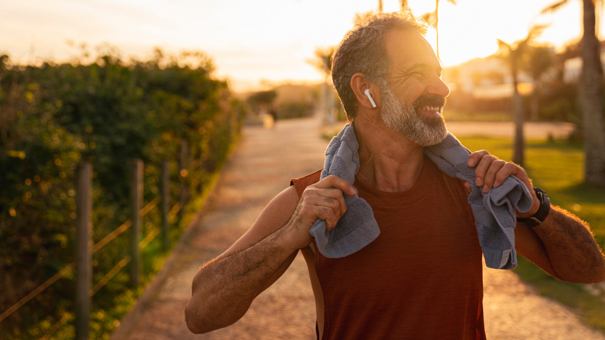 Ein lächelnder älterer Mann, der sich im Freien bei Sonnenuntergang aufhält und kabellose Kopfhörer trägt. Er trägt ein Handtuch um den Hals hält dieses mit beiden Händen fest.