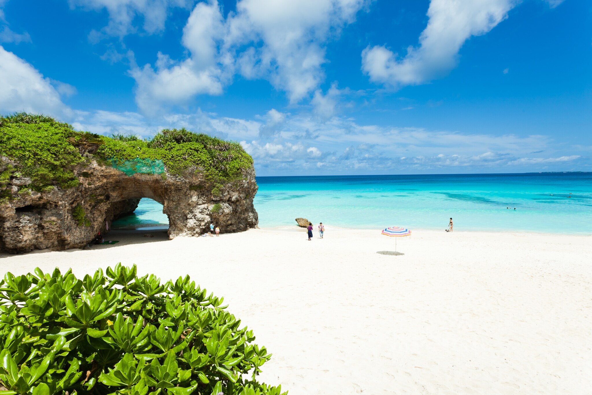 Personen an einem weißen Sandstrand mit bewachsenem Felsen an türkisblauem Meer.