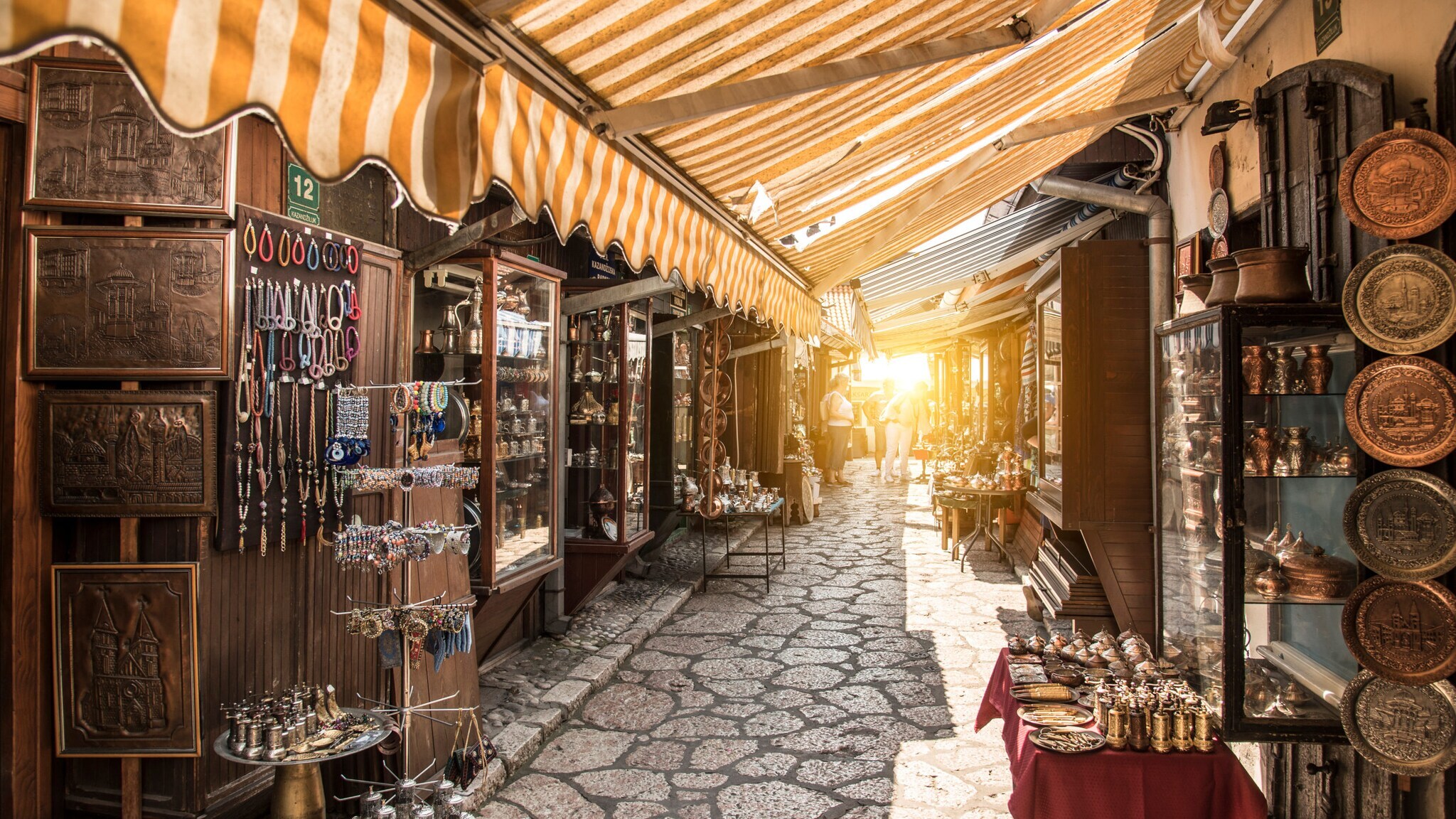 Enger Marktgang mit gepflastertem Boden, beidseitig Stände mit Schmuck, Keramik und anderen Waren unter gelb-weiß gestreiftem Markisenstoff. Enger Marktgang mit gepflastertem Boden, beidseitig Stände mit Schmuck, Keramik und anderen Waren unter gelb-weiß gestreiftem Markisenstoff.