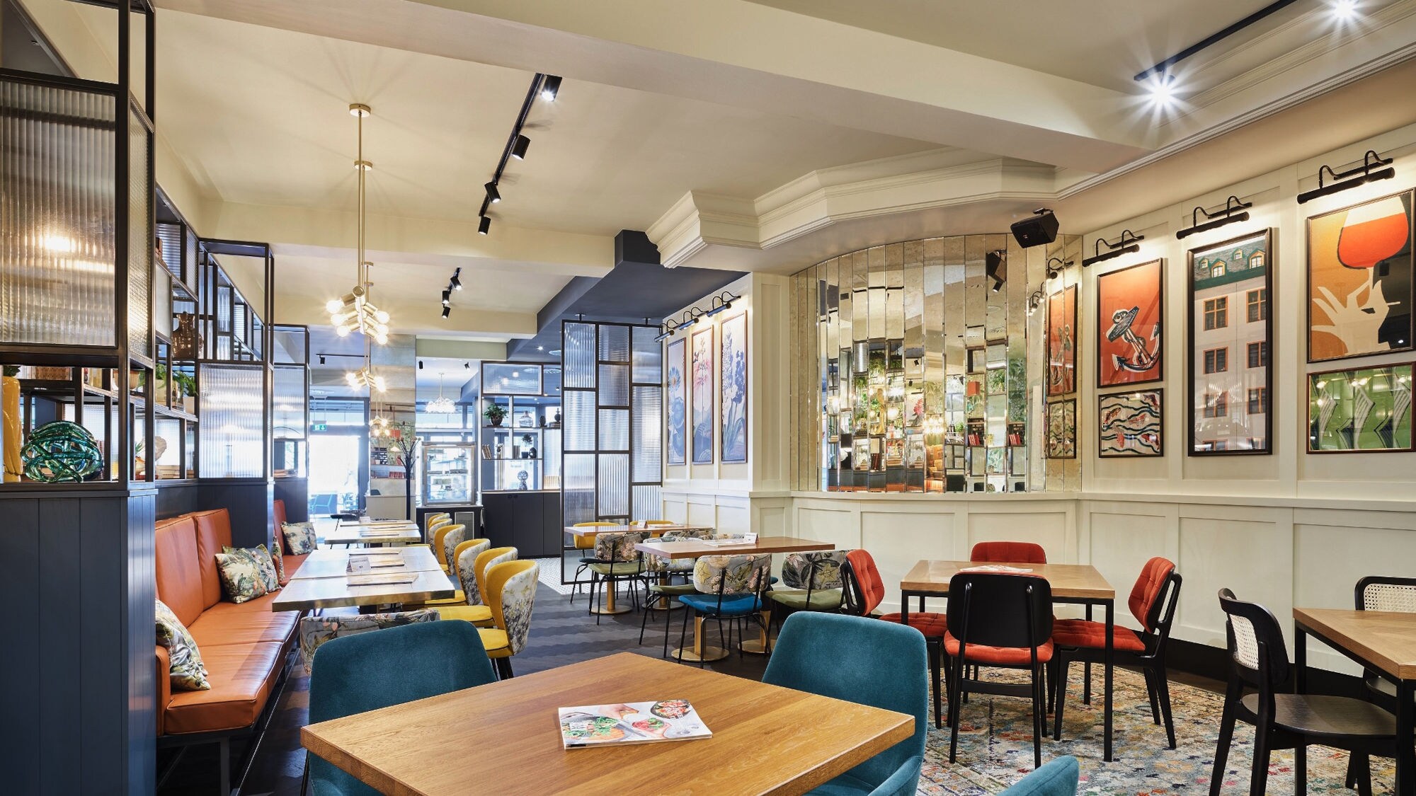 Innenraum eines Cafés mit Holztischen, gepolsterten Stühlen in Blau, Rot und Schwarz, Spiegelwand und moderner Beleuchtung.