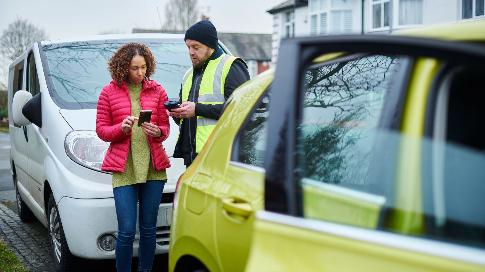 Eine Frau und ein Mann in gelber Warnweste im Austausch miteinander mit Smartphones in den Händen zwischen zwei geparkten Autos in einer Wohnsiedlung. Eine Frau und ein Mann in gelber Warnweste im Austausch miteinander mit Smartphones in den Händen zwischen zwei geparkten Autos in einer Wohnsiedlung.