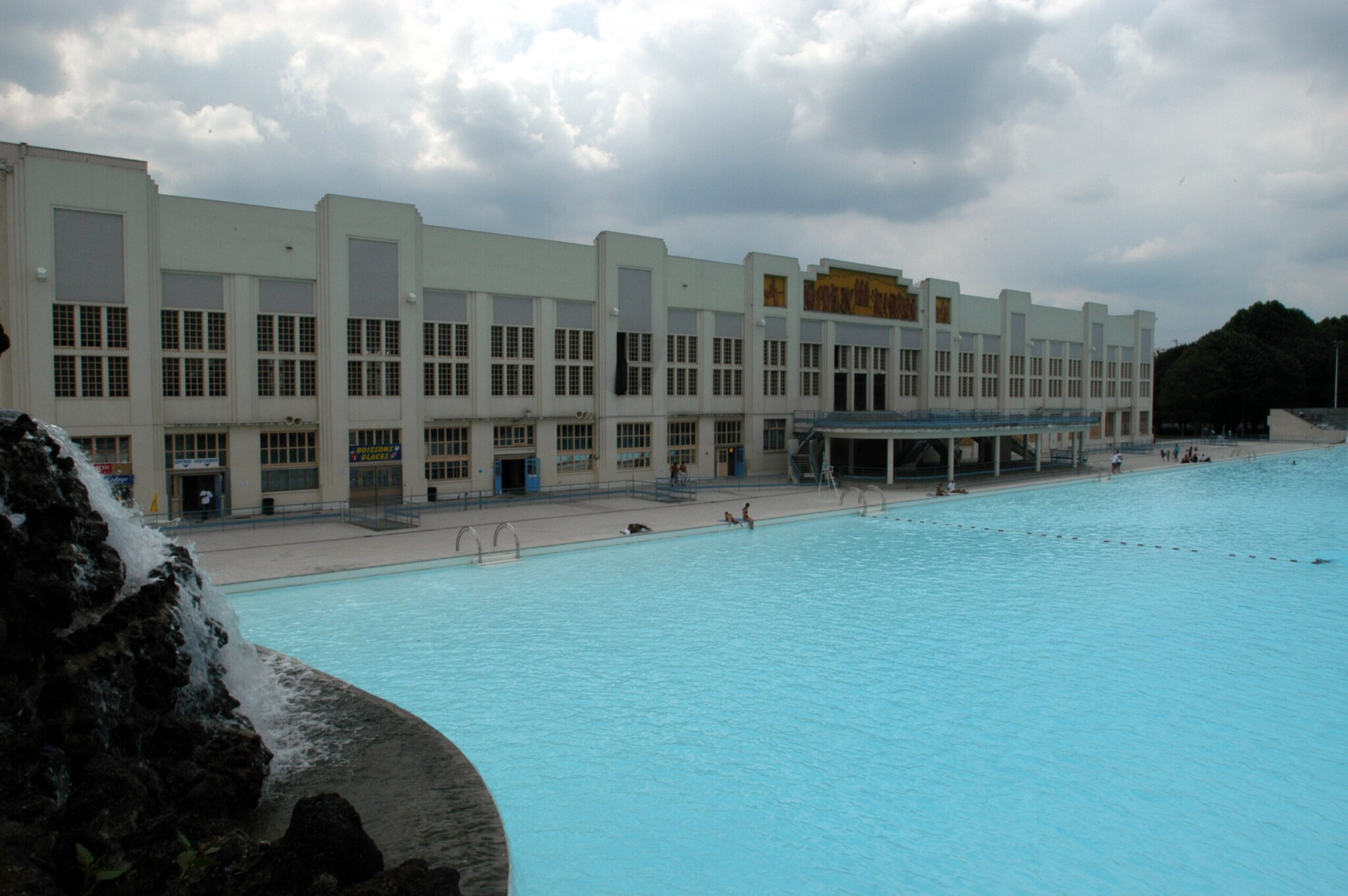 Ein großes blaues Schwimmbecken vor dem Hintergrund eines genauso großen Gebäudes im Art-déco-Stil Ein großes blaues Schwimmbecken vor dem Hintergrund eines genauso großen Gebäudes im Art-déco-Stil