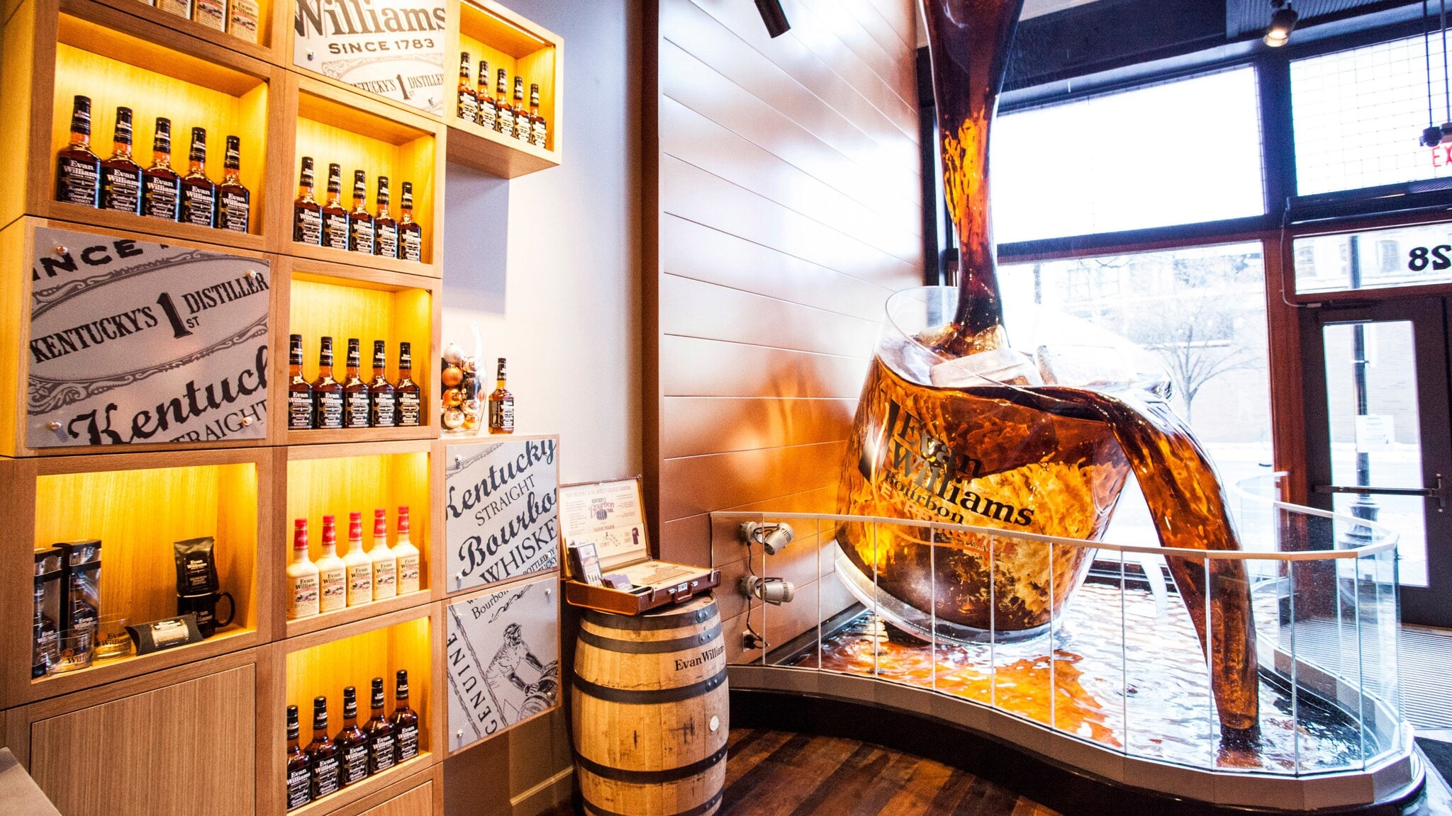 Große Skulptur eines Bourbon-Glases neben Regalen mit Evan Williams Bourbonflaschen und einem Holzfass im Ausstellungsraum Große Skulptur eines Bourbon-Glases neben Regalen mit Evan Williams Bourbonflaschen und einem Holzfass im Ausstellungsraum