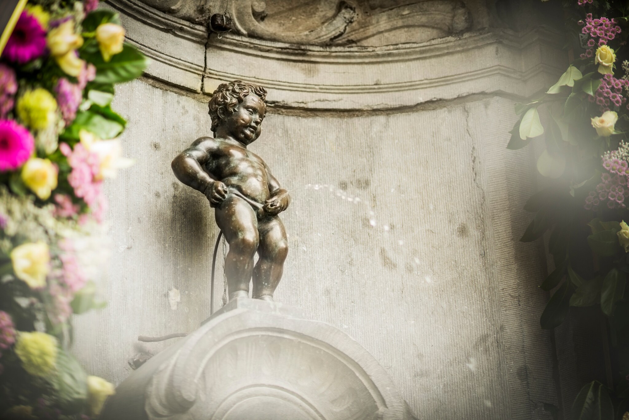 Bronzefigur eines pinkelnden Jungen an einer Fassade, im Vordergrund Blumendekoration.