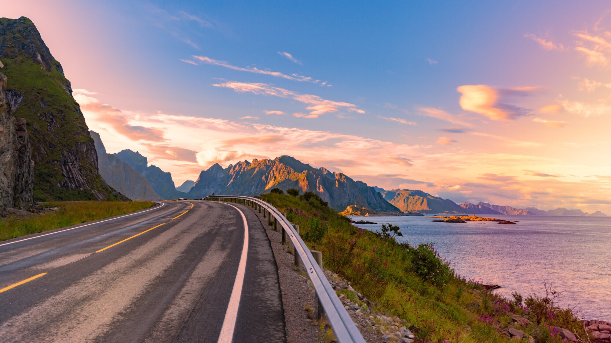 Panorama einer Straße bei Sonnenuntergang auf den norwegischen Lofoten, die am Meer entlangführt. Im Hintergrund Berge.