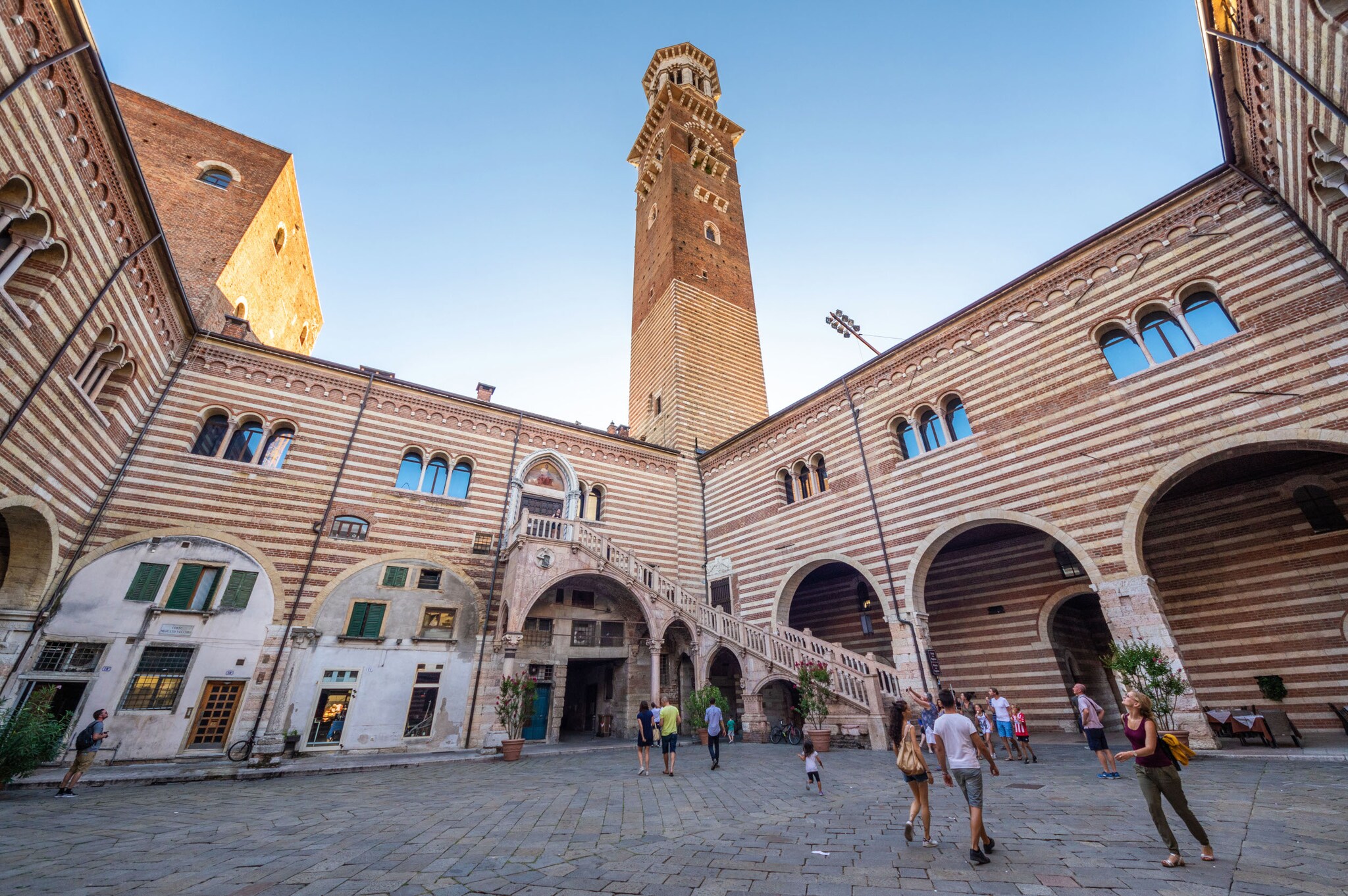 Personen in einem italienischen Palastinnenhof mit gestreifter Fassade und Außentreppe unter einem Turm vor blauem Himmel.