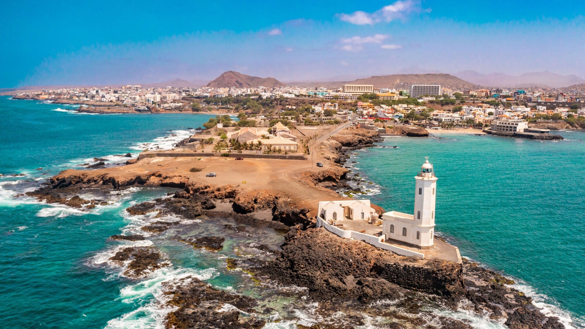 Luftaufnahme eines Leuchtturms auf einer felsigen Küste, umgeben von türkisblauem Wasser und Wellen, mit einer Stadt im Hintergrund. Luftaufnahme eines Leuchtturms auf einer felsigen Küste, umgeben von türkisblauem Wasser und Wellen, mit einer Stadt im Hintergrund.