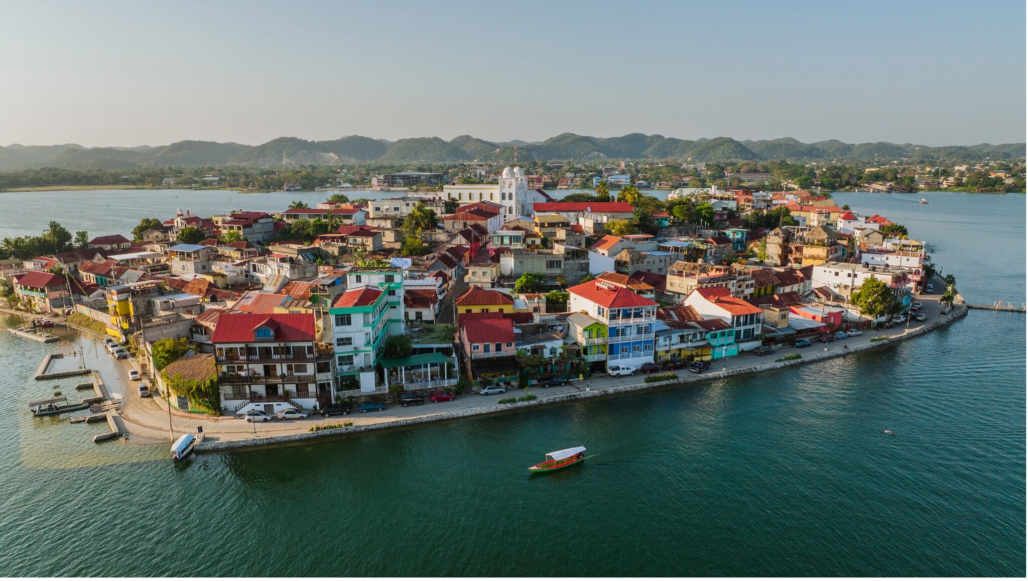 Luftaufnahme einer kleinen Insel mit dicht bebauten Häusern mit roten Dächern, umgeben von Wasser und Bergen im Hintergrund