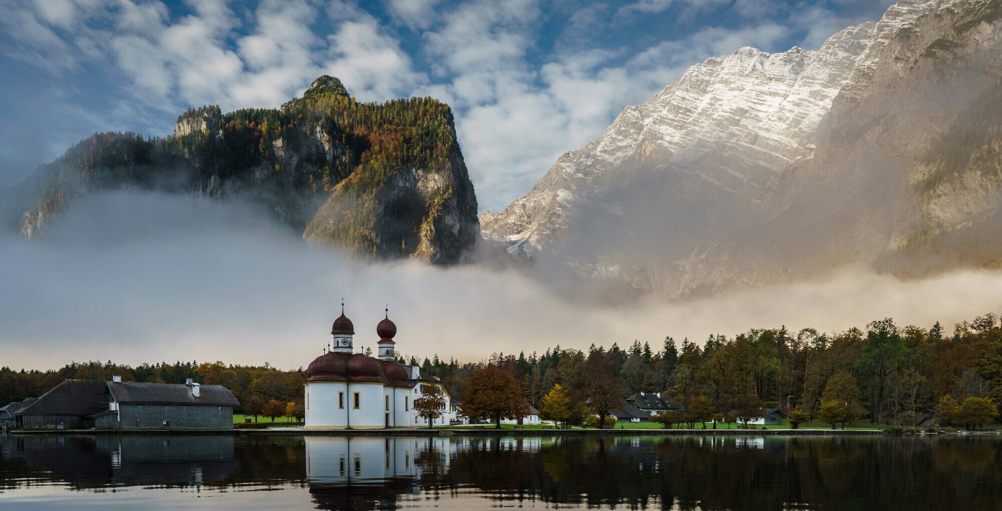 Eine weiße Kapelle mit roten Zwiebeltürmen in der herbstlichen Landschaft am Ufer eines Sees vor zwei riesigen Felswänden