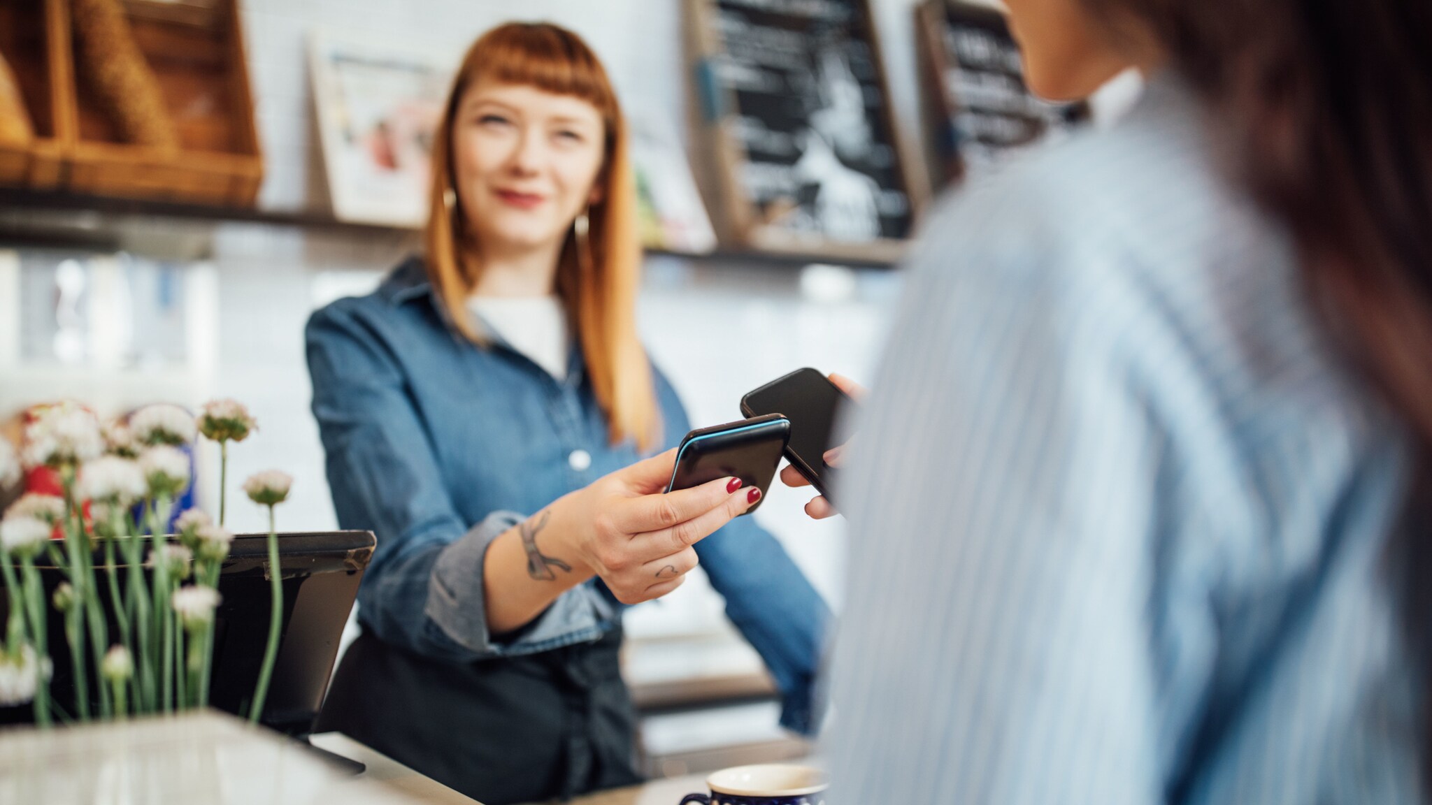 Kontaktlose Zahlung per Smartphone an einem Kartenlesegerät, gehalten von einer Frau hinter einer Theke in einem Geschäft.