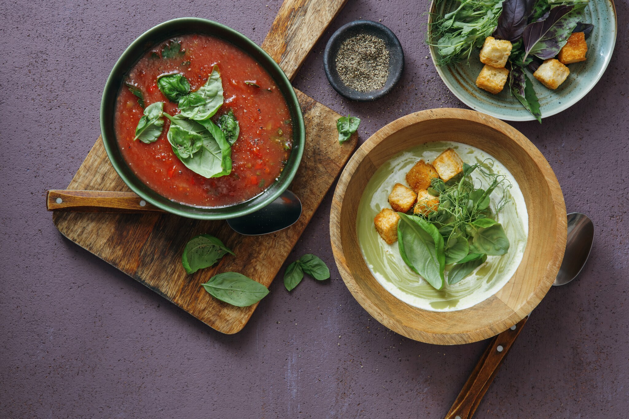 Aufsicht einer roten und einer grünen Gemüsesuppe, garniert mit Croutons und Kräutern, in Schalen auf einem Tisch