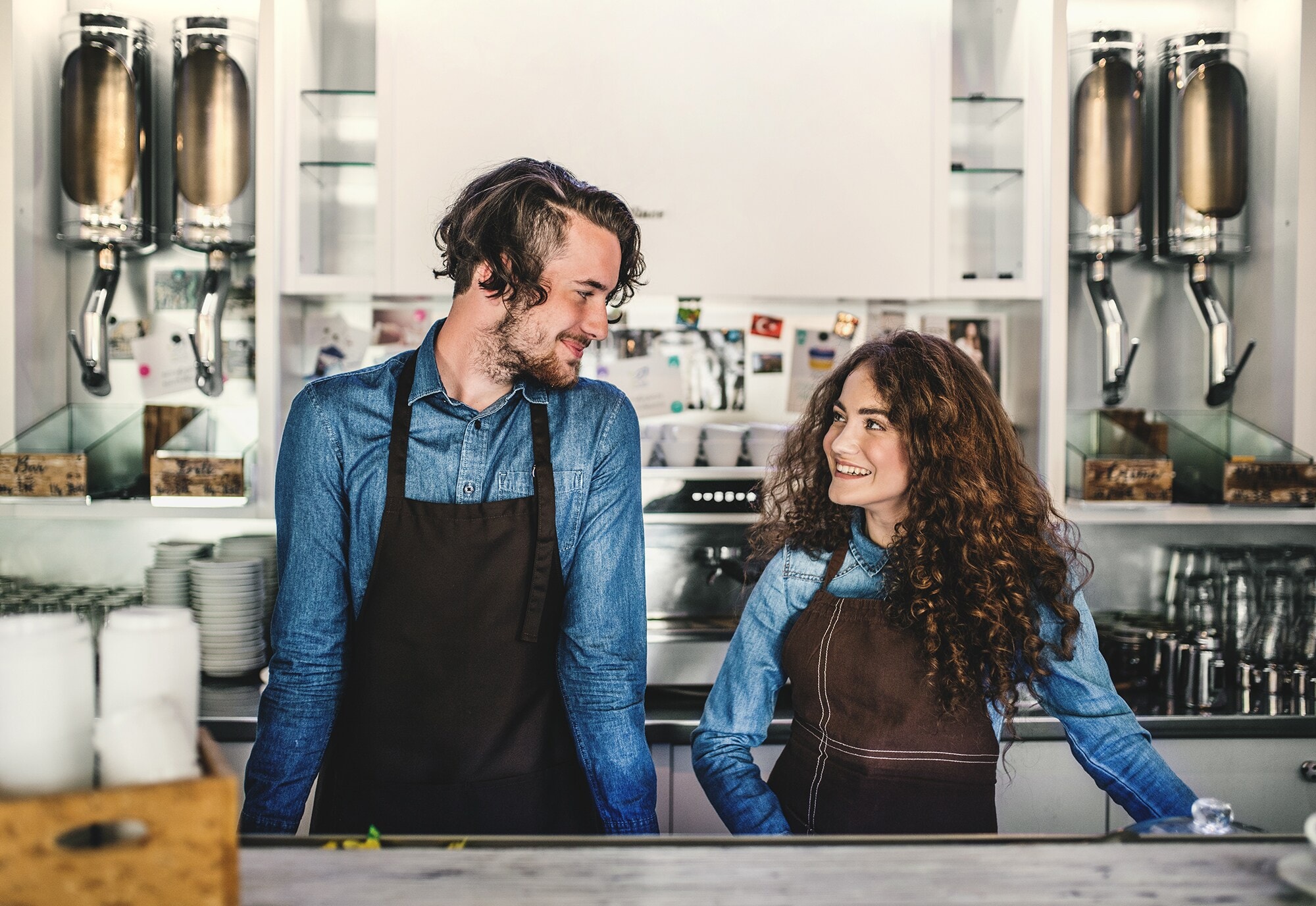 Gute Freund:innen – gute Geschäftspartner:innen? So beginnt eure Erfolgsstory Zwei junge Menschen in Jeanshemd mit einer Schürze stehen nebeneinander an einem Tresen vor einer Siebträgermaschine und Kaffeemühlen.