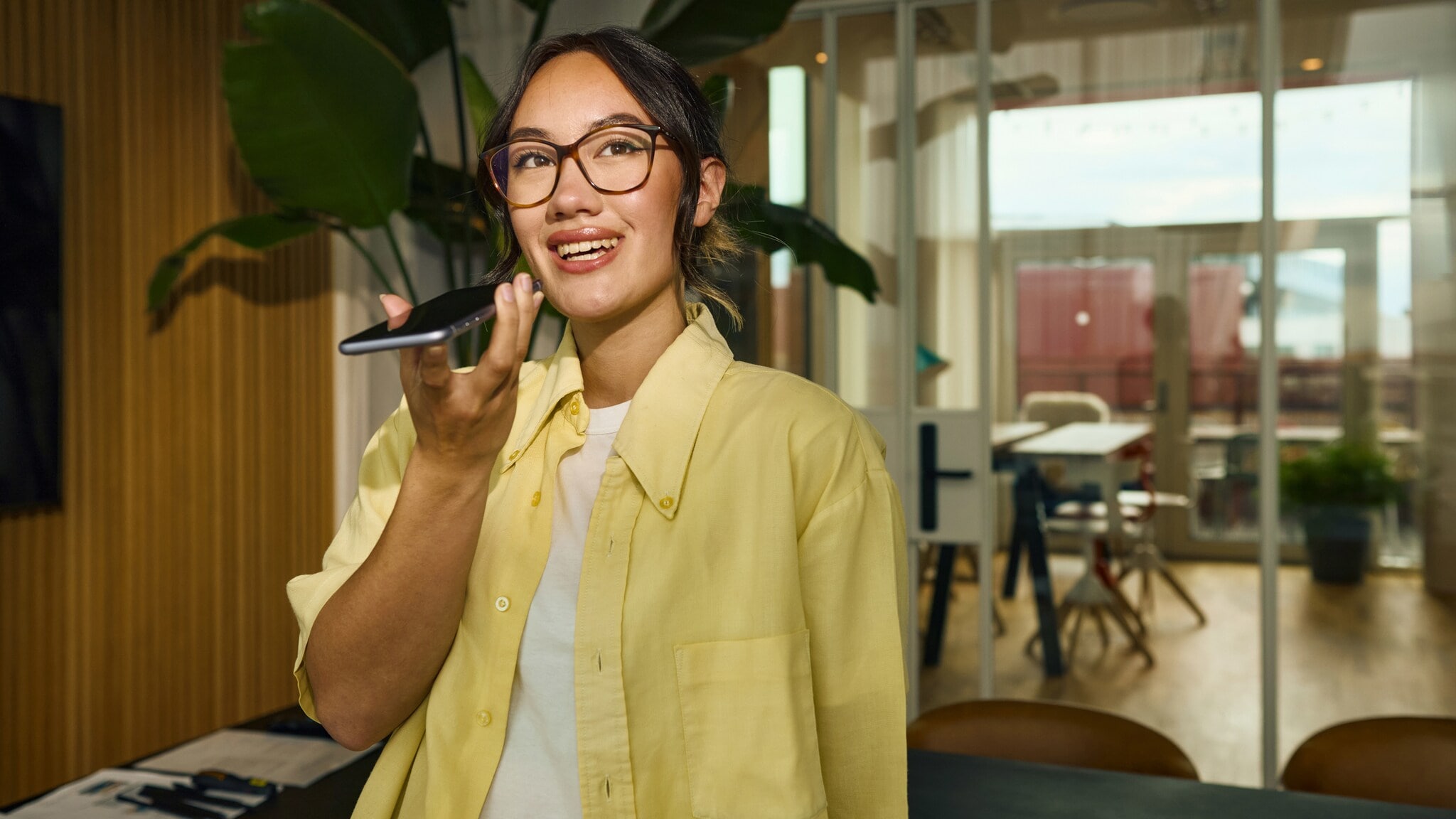 Eine junge Frau im gelben Hemd spricht in ein Smartphone in einem Büro. Eine junge Frau im gelben Hemd spricht in ein Smartphone in einem Büro.