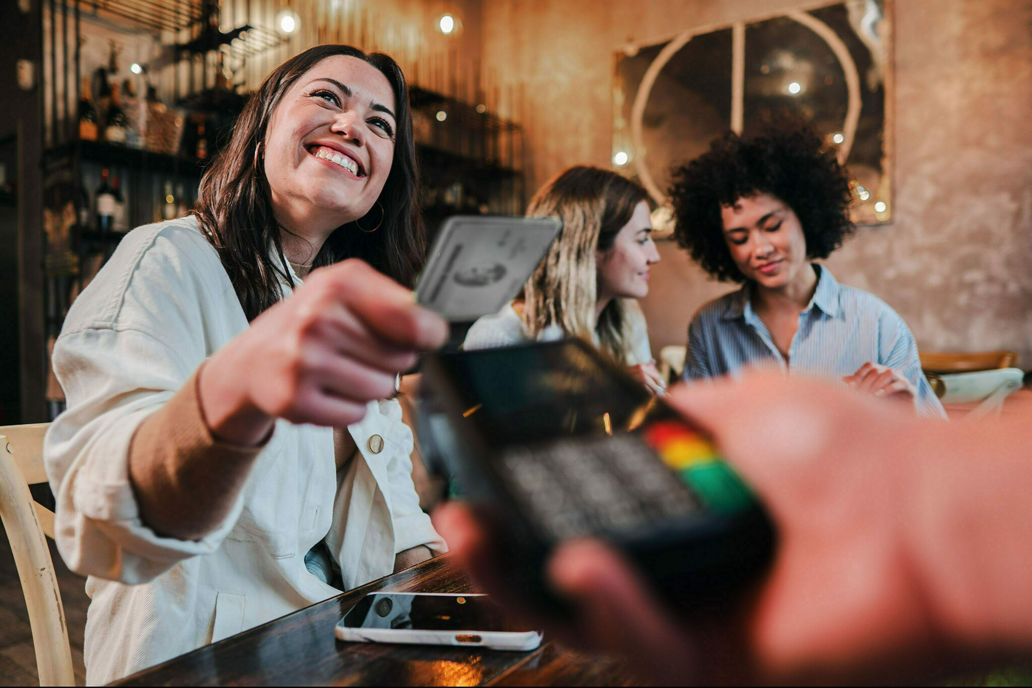 Eine junge Frau bezahlt kontaktlos mit einer silbernen Kreditkarte an einem Kartenlesegerät in einem Restaurant.