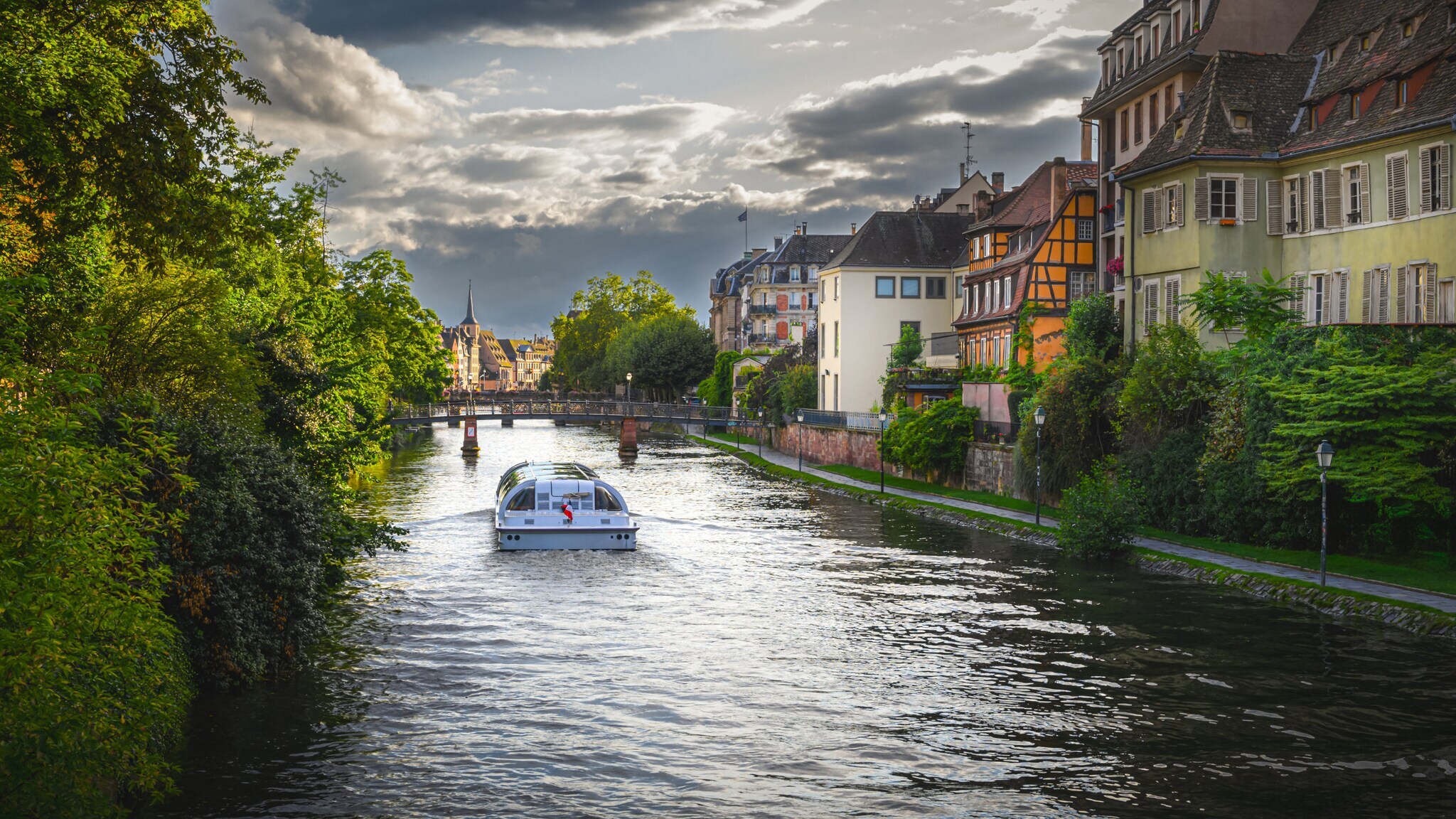 Boot fährt auf einem Fluss zwischen grünen Bäumen und Fachwerkhäusern in einer europäischen Stadt Boot fährt auf einem Fluss zwischen grünen Bäumen und Fachwerkhäusern in einer europäischen Stadt