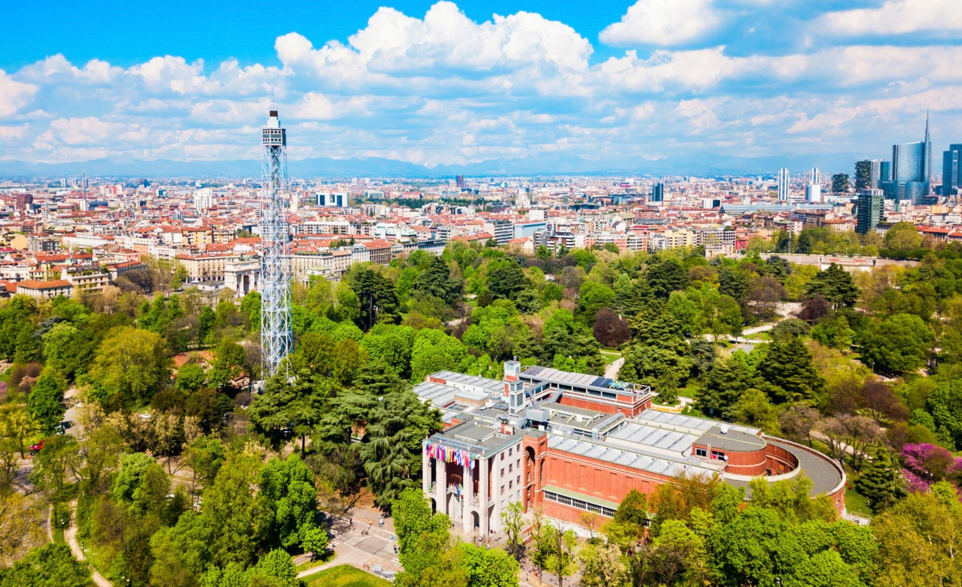 Blick von oben auf einen grünen Park mit einem großen Gebäude und einem hohen Stahlturm in der Mitte, im Hintergrund die Häuser einer Stadt. Blick von oben auf einen grünen Park mit einem großen Gebäude und einem hohen Stahlturm in der Mitte, im Hintergrund die Häuser einer Stadt.