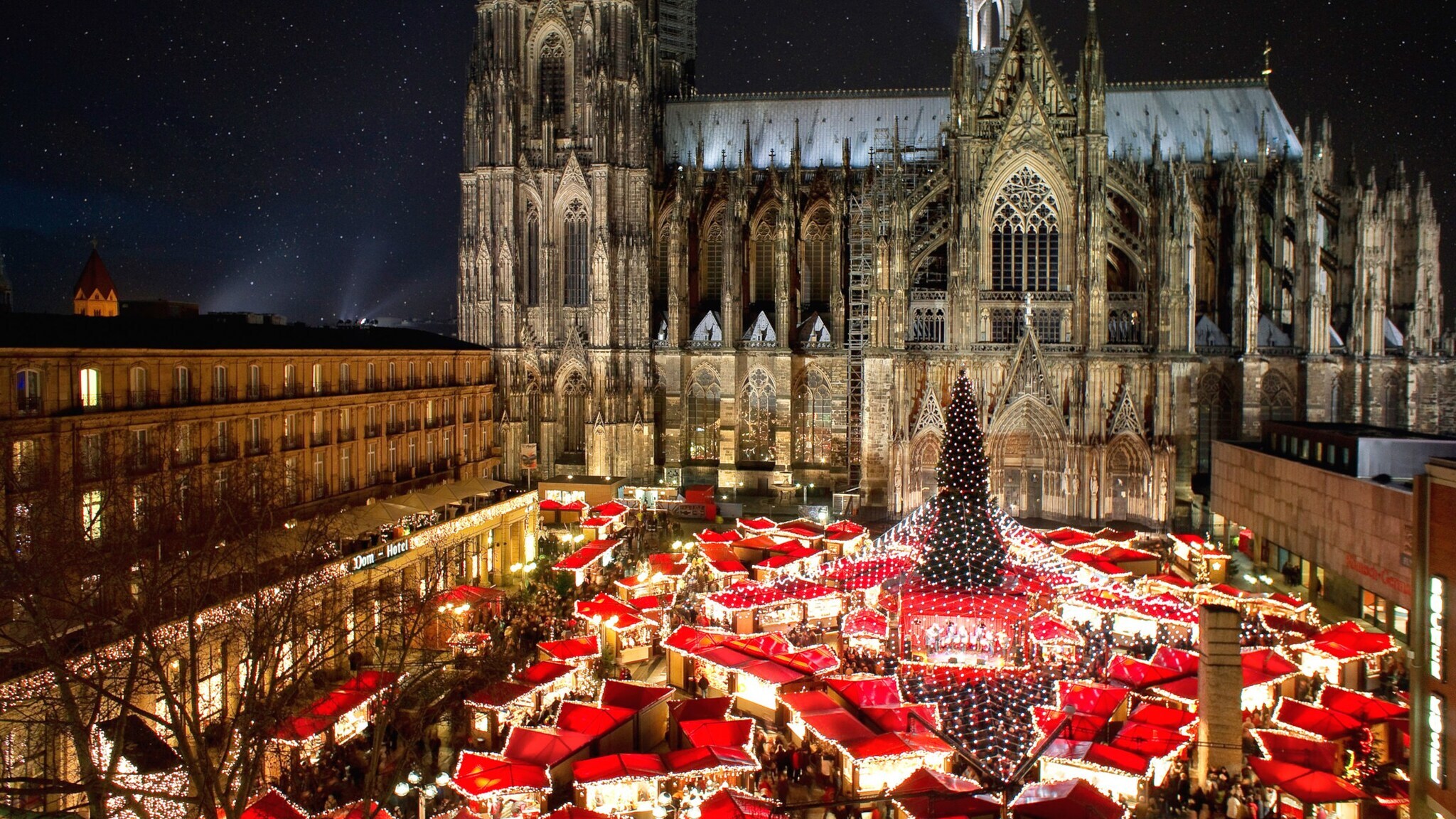 Beleuchteter Weihnachtsmarkt mit Tannenbaum am Kölner Dom bei Dunkelheit.