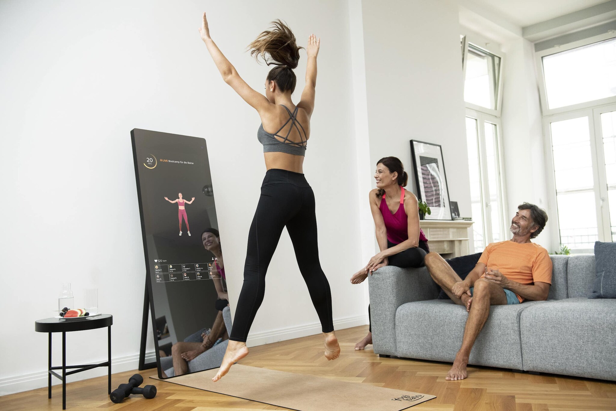 Eine junge Frau in Sportkleidung springt vor einem interaktiven Spiegel in die Luft, neben ihr sitzen auf einem grauen Sofa ein Mann und eine Frau, ebenfalls in Sportkleidung, links im Bild steht noch ein kleiner Tisch mit Obst und Wassergläsern und auf dem Boden liegen schwarze Hanteln