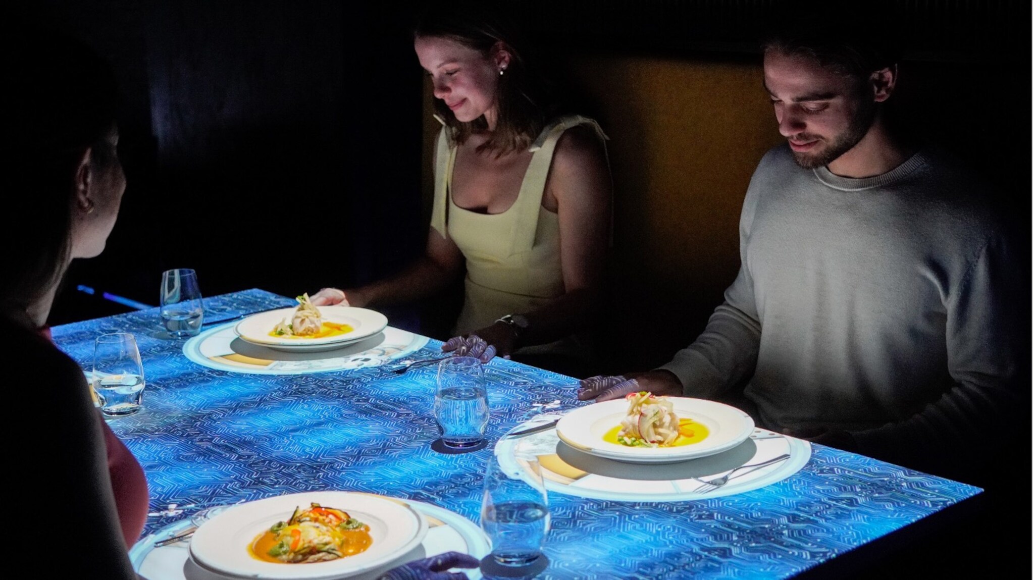 Drei Personen sitzen an einem Tisch mit Projektion. Auf dem Tisch sind zwei Teller mit einer Speise und Gläser sichtbar. Der Tisch hat ein blaues, gemustertes Design