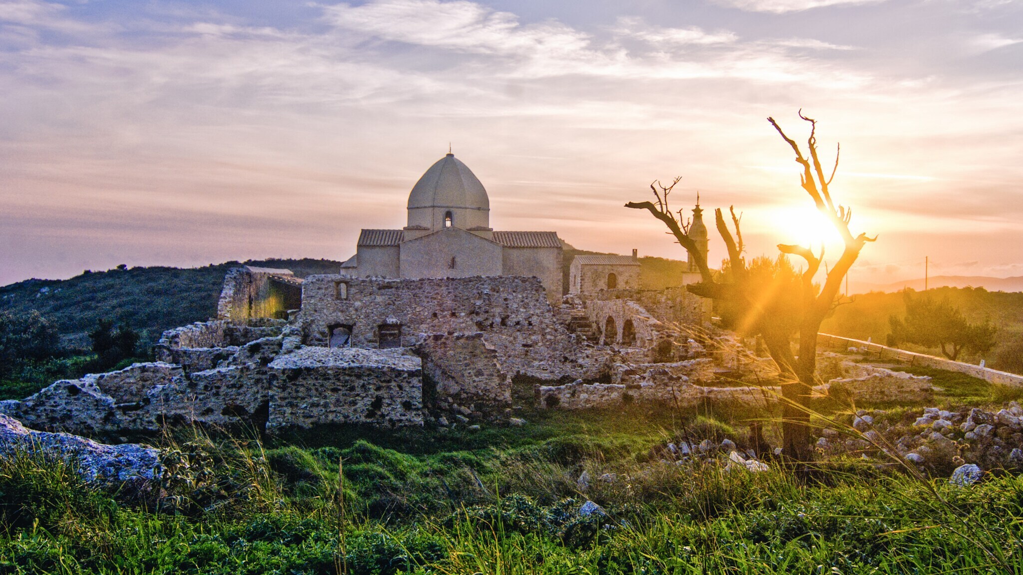 Sonnenuntergang über einer alten Steinruine mit einer Kuppel und einem kahlen Baum im Vordergrund