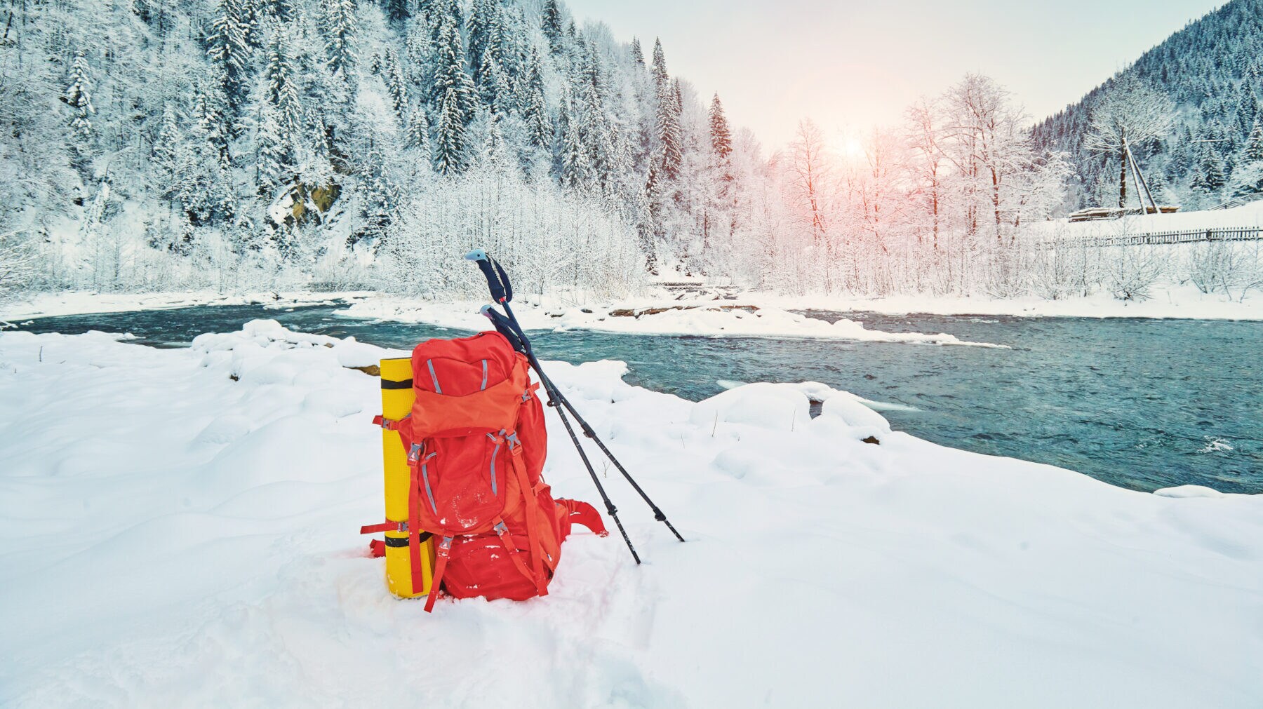 An einem Fluss steht ein Rucksack mit an ihm befestigter Matte und angelehnten Wanderstöcken im Schnee