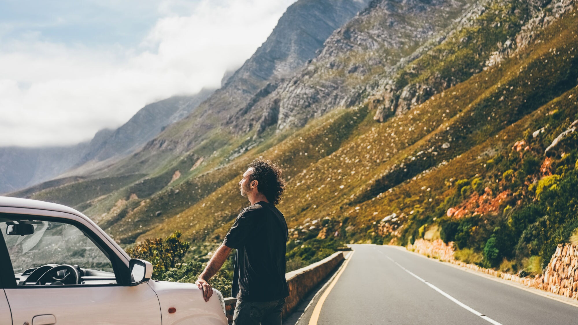 Mann lehnt an weißem Auto an einer Bergstraße, umgeben von steilen Felsen und bewölktem Himmel.