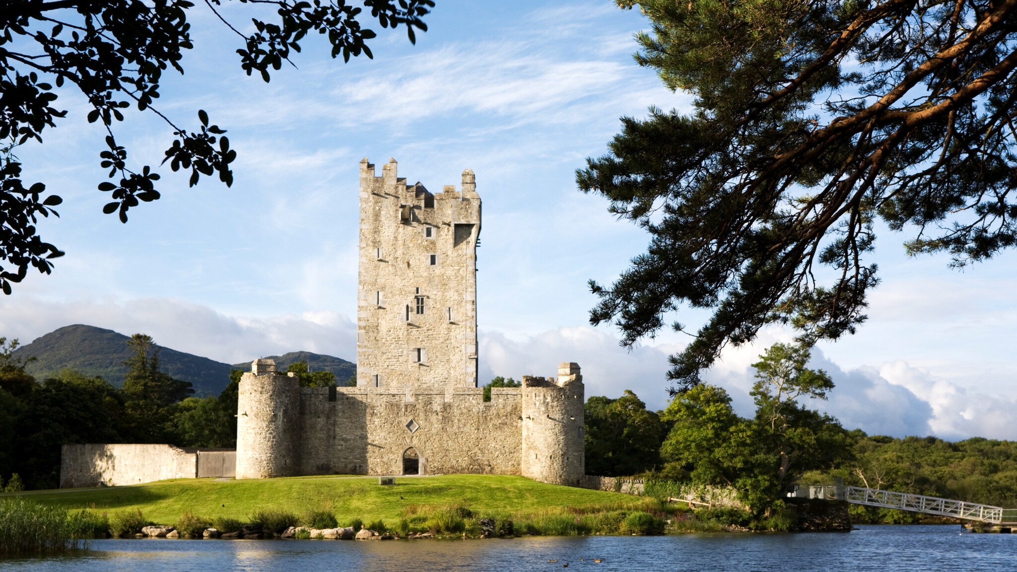 Eine mittelalterliche Burg mit hohen Türmen und dicken Mauern, umgeben von Bäumen und Wasser. Im Hintergrund sind sanfte Hügel sichtbar. Eine mittelalterliche Burg mit hohen Türmen und dicken Mauern, umgeben von Bäumen und Wasser. Im Hintergrund sind sanfte Hügel sichtbar.