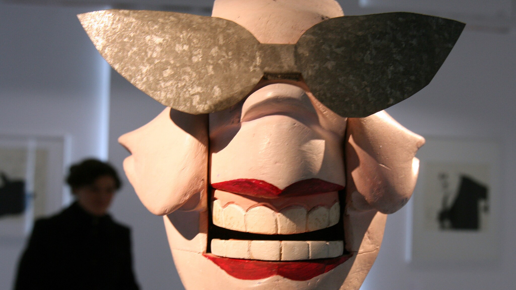 Große, stilisierte Gesichtsmaske mit übergroßen Metallsonnenbrillen und roten Lippen, ausgestellt in einer Galerie Große, stilisierte Gesichtsmaske mit übergroßen Metallsonnenbrillen und roten Lippen, ausgestellt in einer Galerie