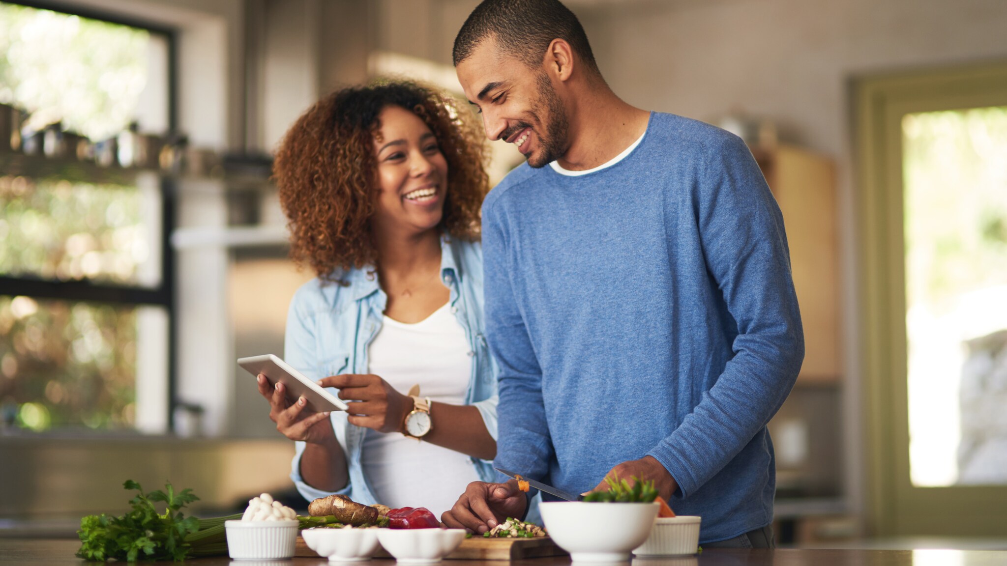 Zwei Personen stehen in einer Küche, eine hält ein Tablet, auf dem Tisch liegen verschiedene Schüsseln mit Lebensmitteln.
