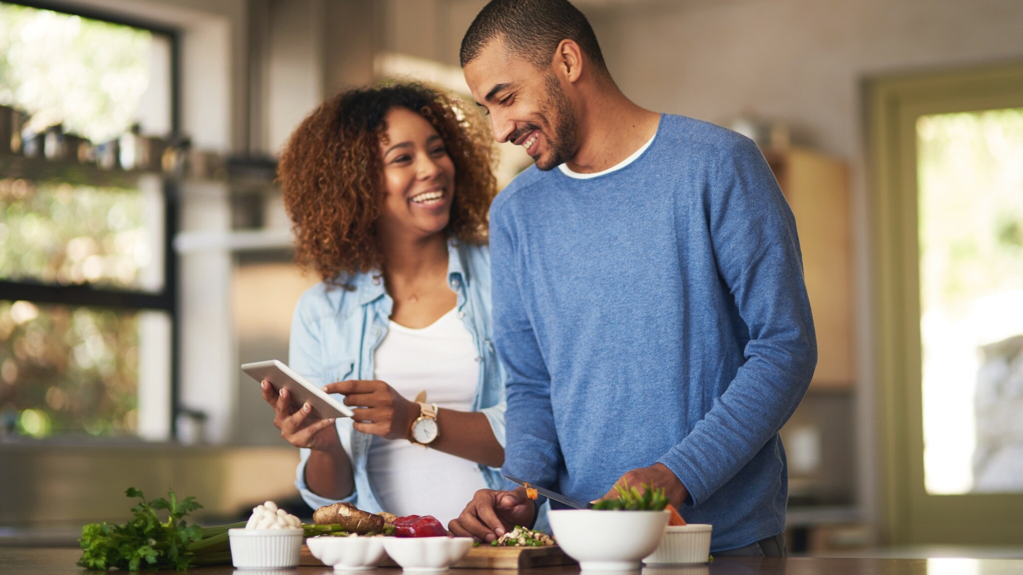 Zwei Personen stehen in einer Küche, eine hält ein Tablet, auf dem Tisch liegen verschiedene Schüsseln mit Lebensmitteln. Zwei Personen stehen in einer Küche, eine hält ein Tablet, auf dem Tisch liegen verschiedene Schüsseln mit Lebensmitteln.