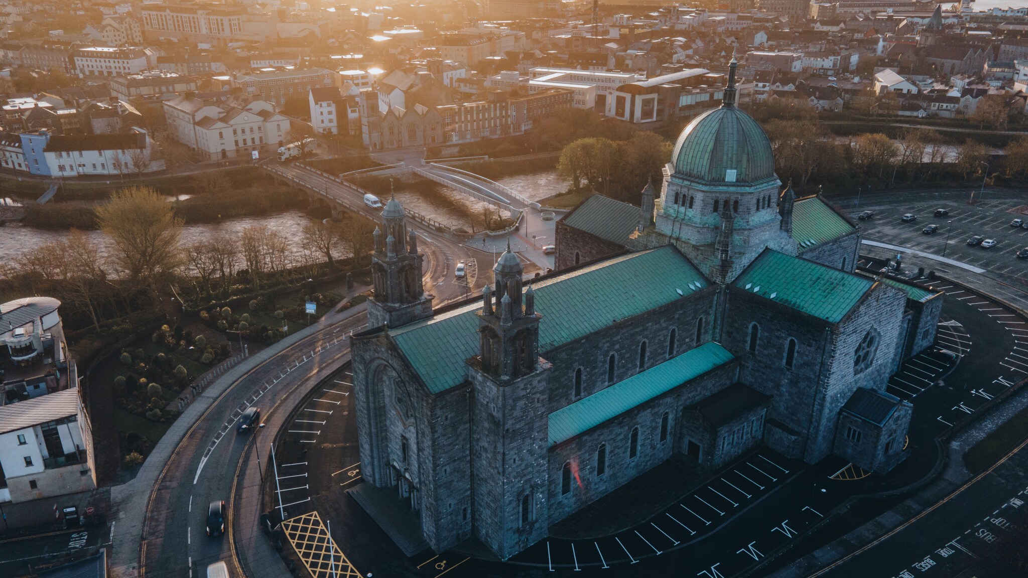 Luftaufnahme einer großen steinernen Kirche mit grüner Kuppel und Türmen, umgeben von einer Stadtlandschaft und Parkplätzen.