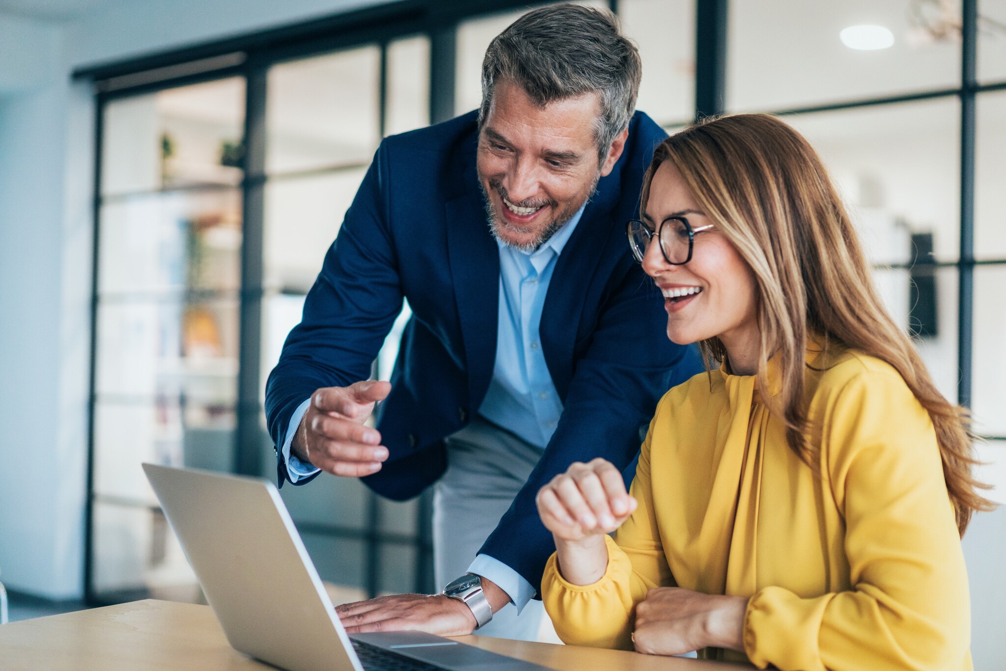 Eine Frau sitzt an einem Schreibtisch, neben ihr steht ein Mann im blauen Sakko, gemeinsam schauen sie lächelnd auf ein Laptop in einem Büroraum.